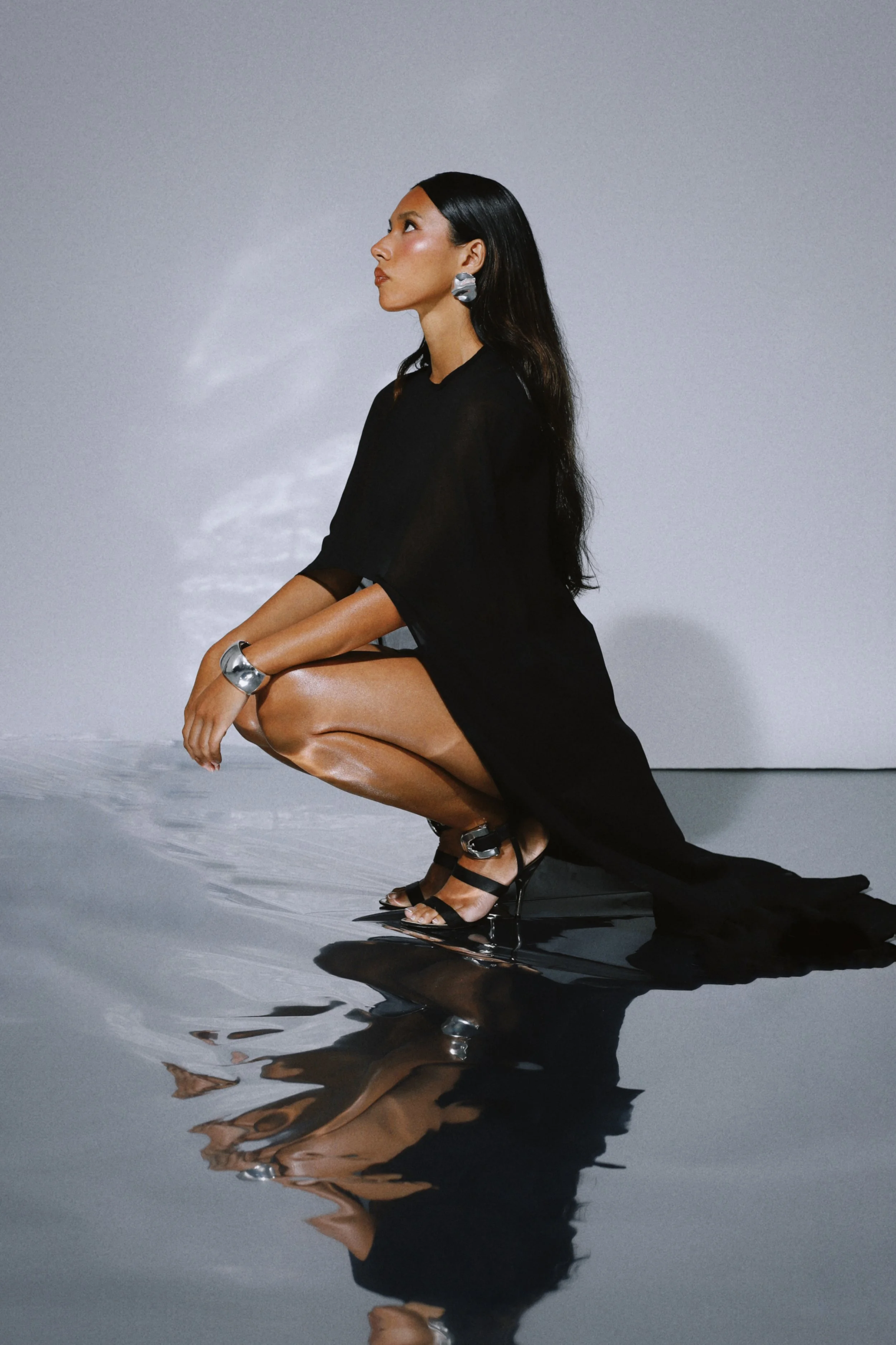 Black sheer kaftan-style dress, model crouched on silver reflective floor. Shot at Style Strategist Studio in Bermondsey, London