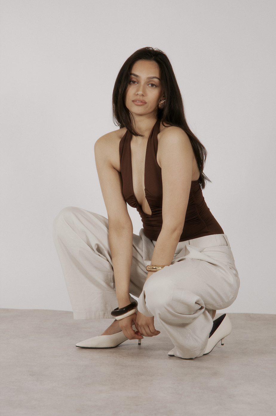 A woman with long dark hair wearing a brown halter top, cream-colored pants, white heeled shoes, and gold jewelry, squatting on the floor against a plain light gray background.