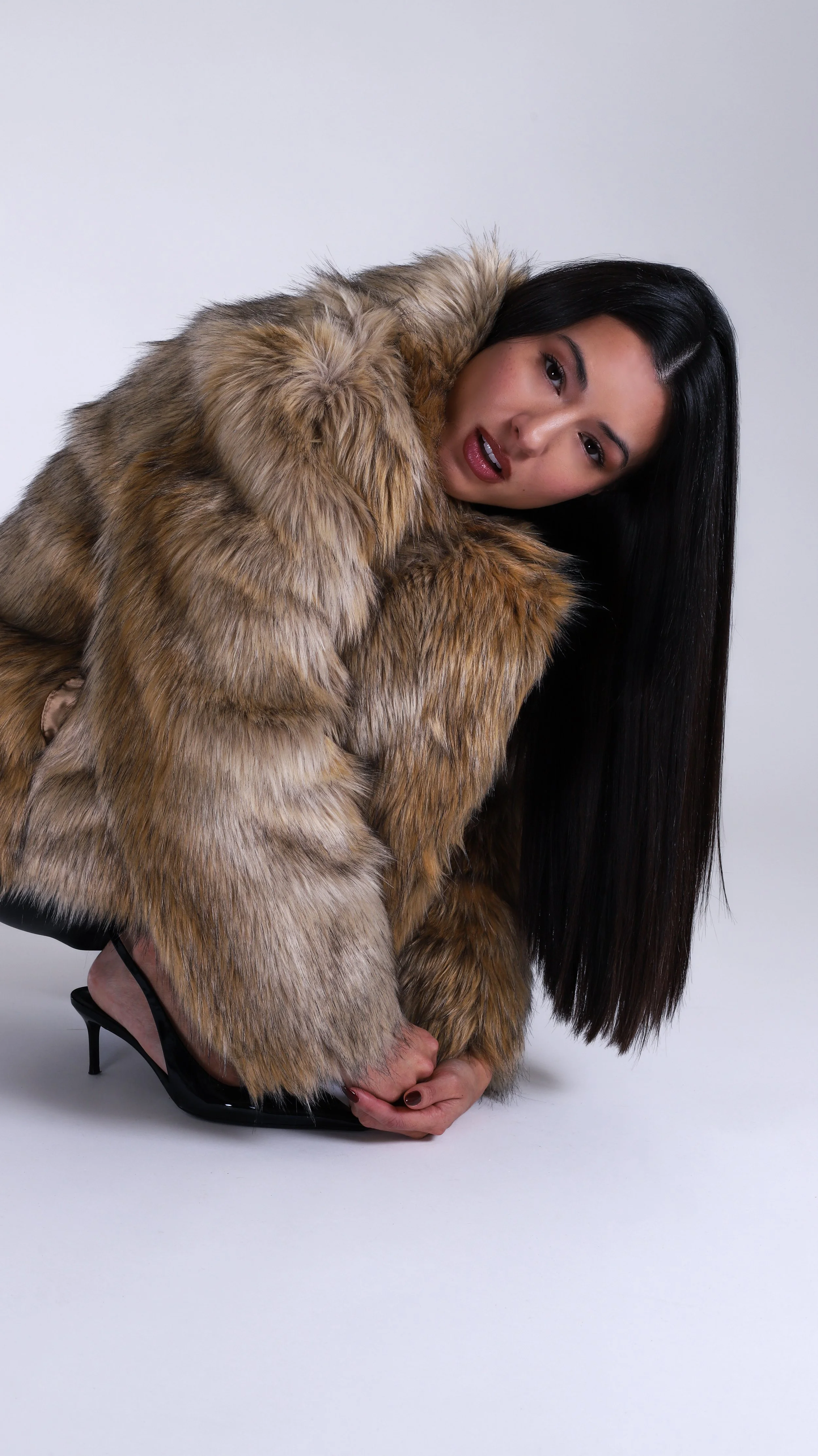 Woman with long black hair wearing a fur coat and high heels, crouching on the floor.
