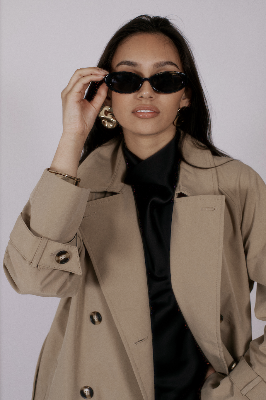 A woman wearing sunglasses, gold earrings, a beige trench coat, and a black shirt poses against a plain background.