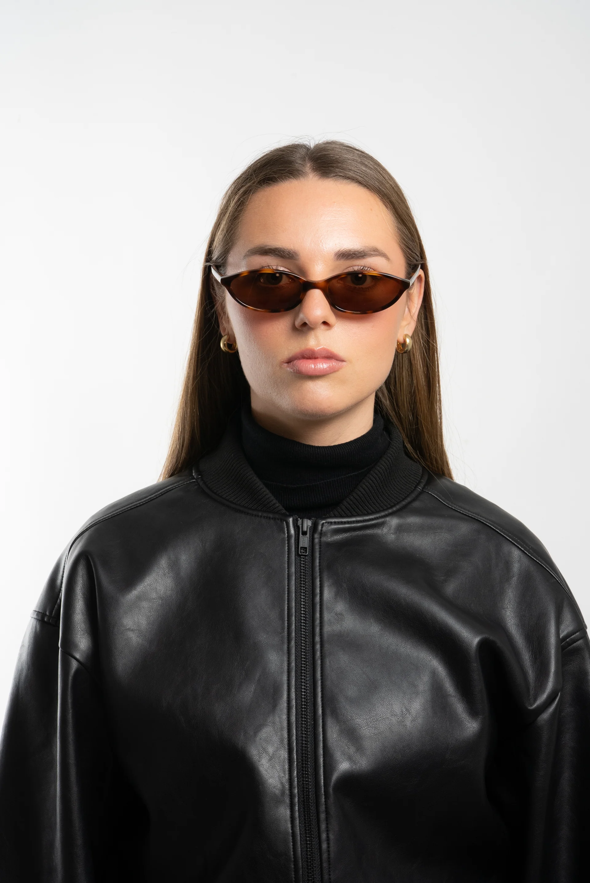A young woman wearing dark sunglasses, a black leather jacket, and gold hoop earrings, against a plain white background.