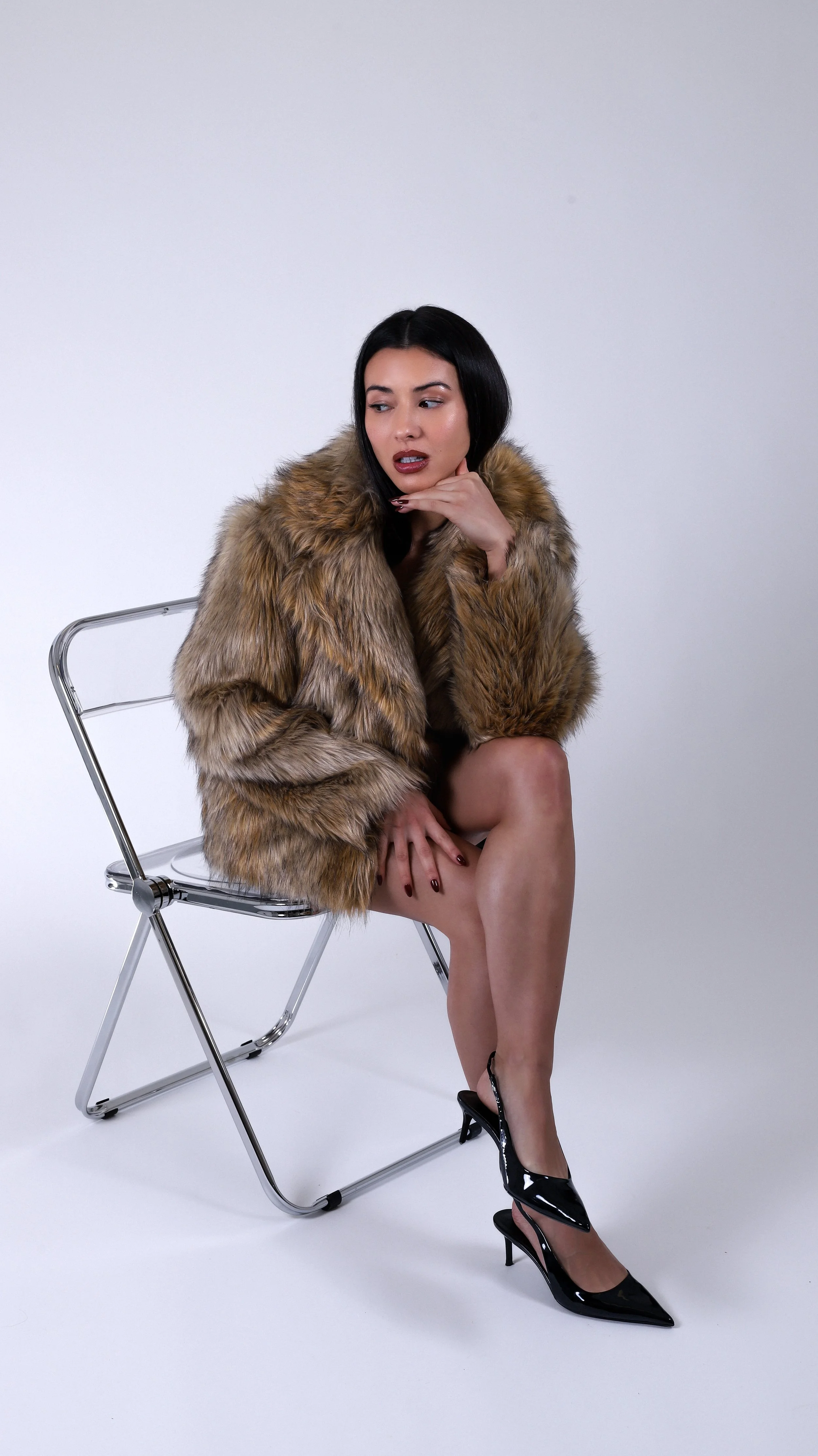 Woman wearing a fur coat and high heels sitting on a metal chair with one leg crossed over the other.