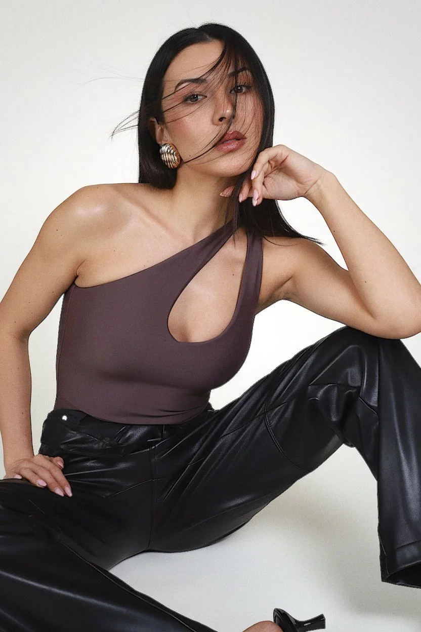 A woman with long dark hair wearing a one-shoulder mauve top with a cutout, large gold earrings, and black leather pants, sitting against a white background.