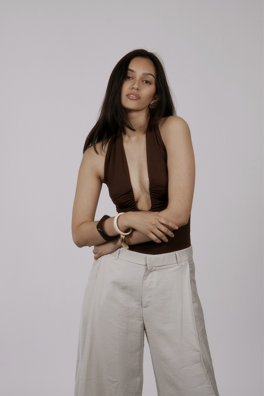 A woman with long dark hair wearing a brown sleeveless top with a deep neckline and light-colored wide-leg pants, posing against a plain light grey background.