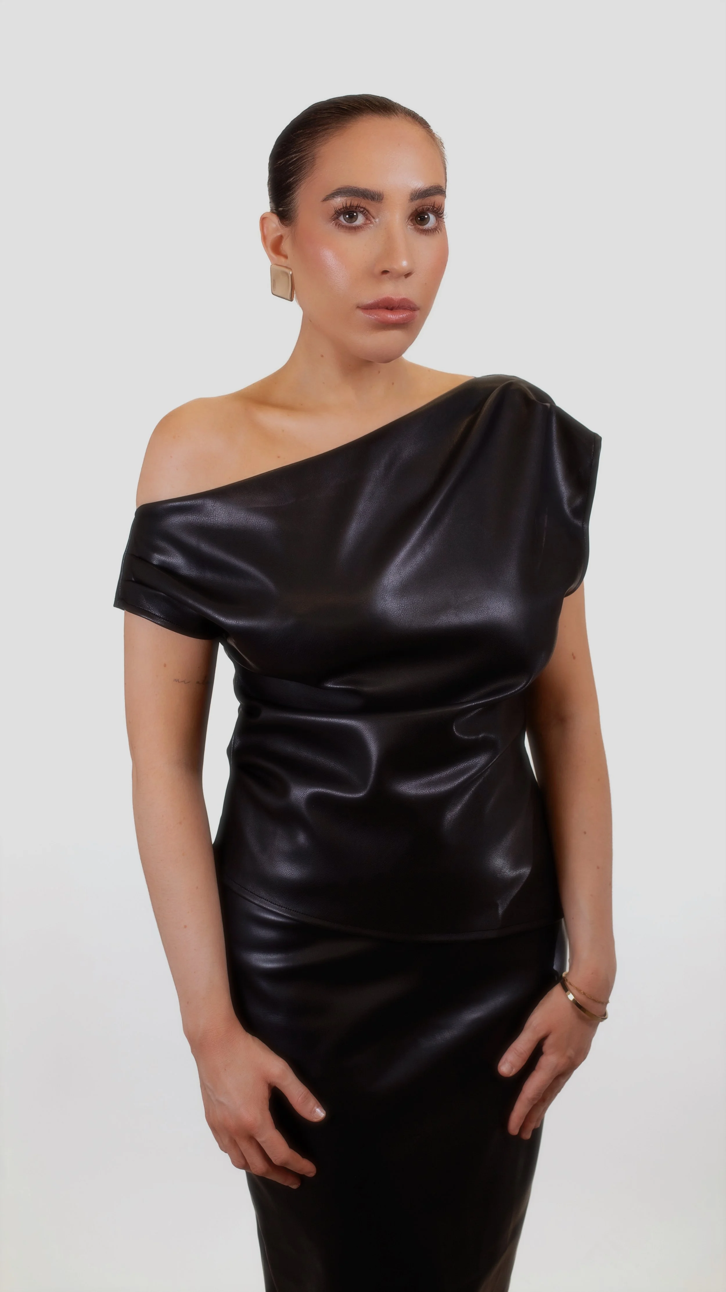 A woman wearing a black leather off-shoulder top and matching skirt, posing against a plain white background.