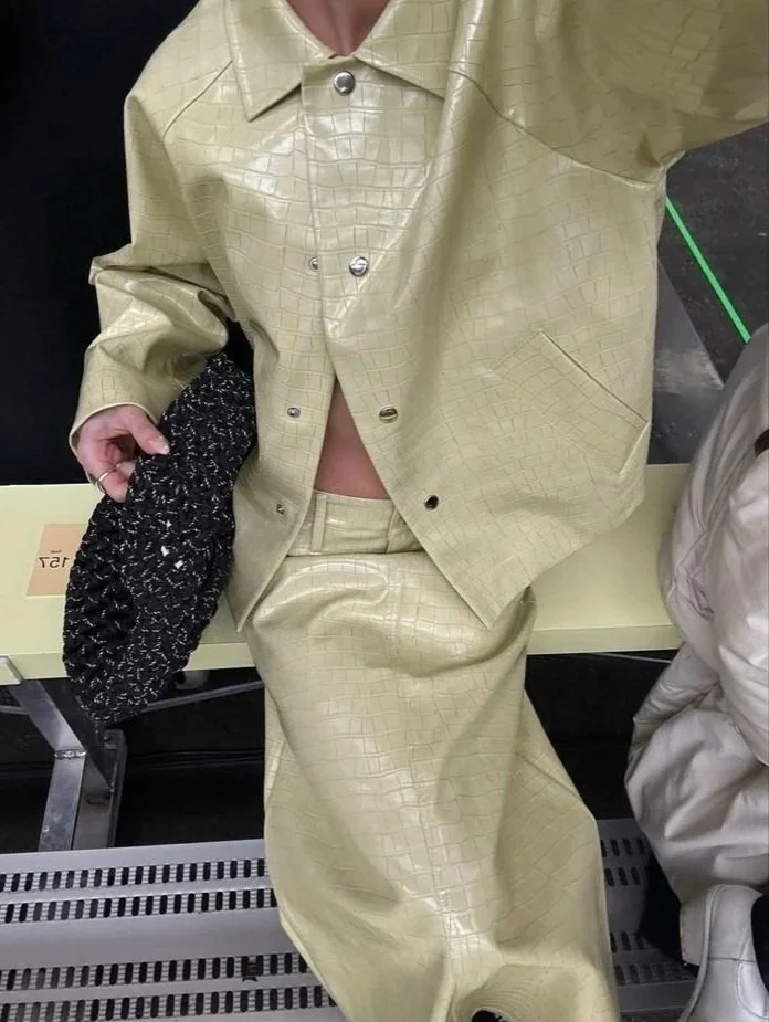 Person wearing a beige, textured jacket and skirt, sitting on a bench, holding a black and white textured bag.