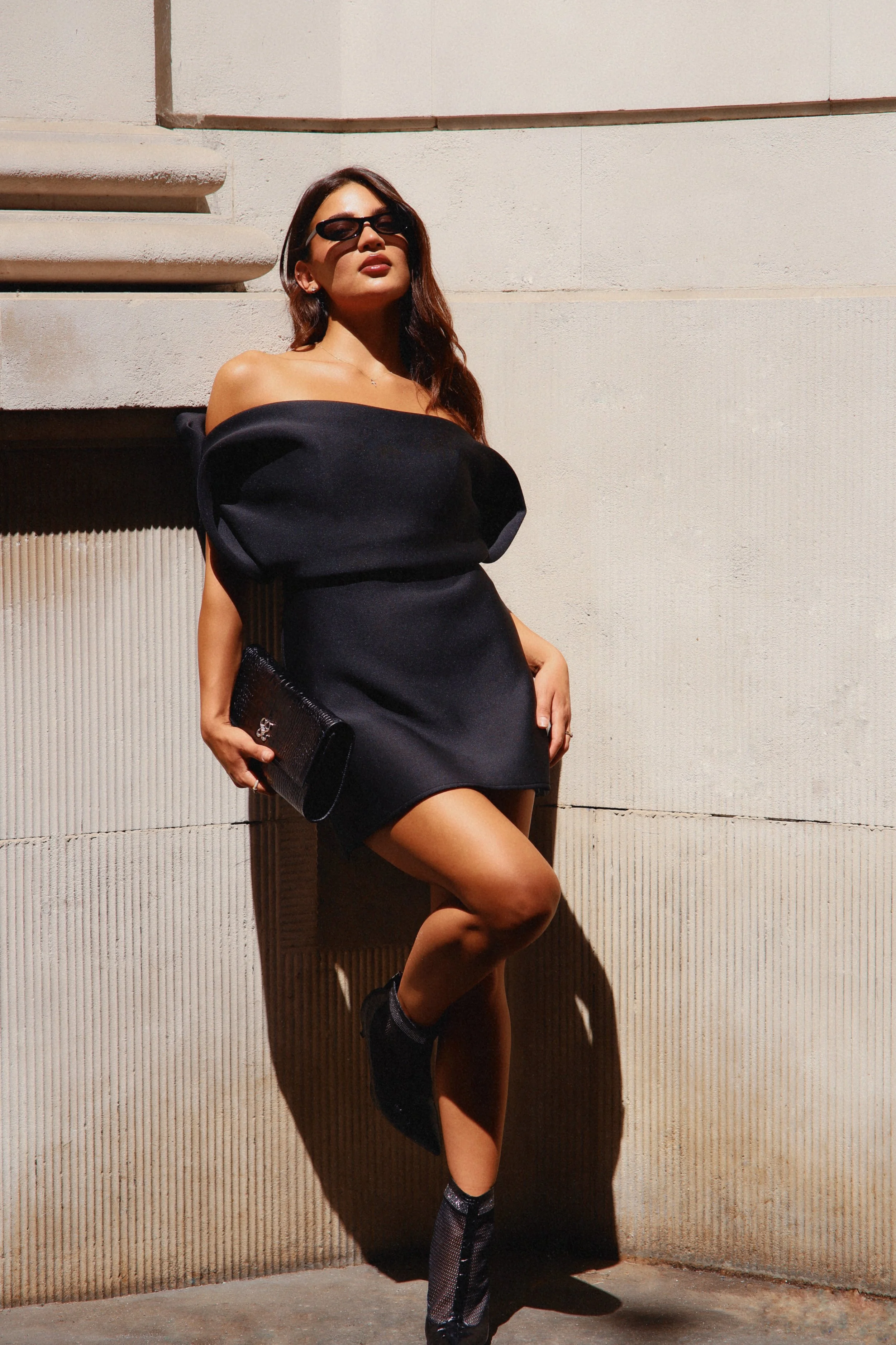 A woman wearing a black off-shoulder dress, black sunglasses, and holding a black clutch, standing against a beige wall with shadow cast on her face and body.