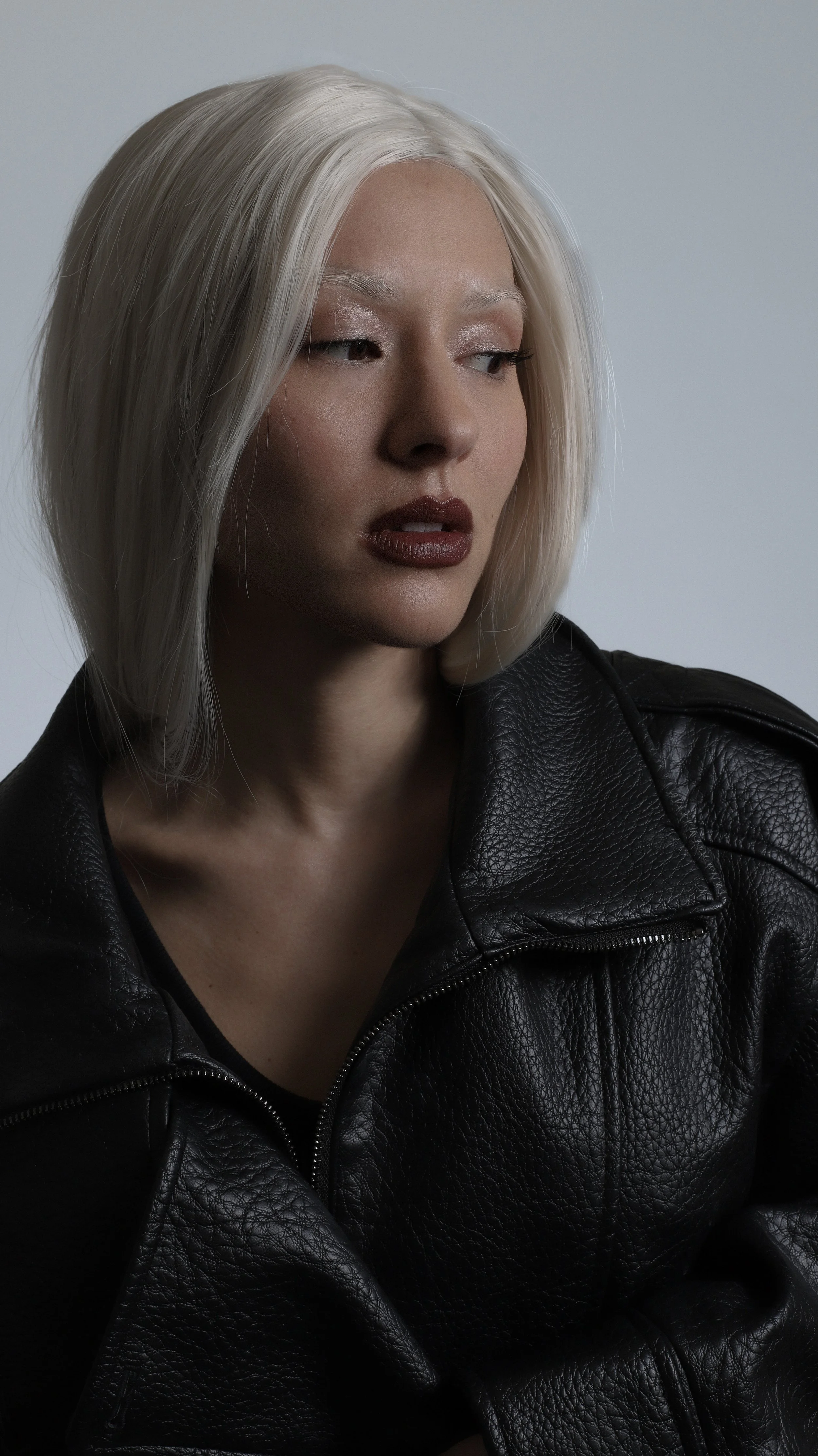A woman with platinum blonde hair wearing a black leather jacket looks to her right against a plain gray background.