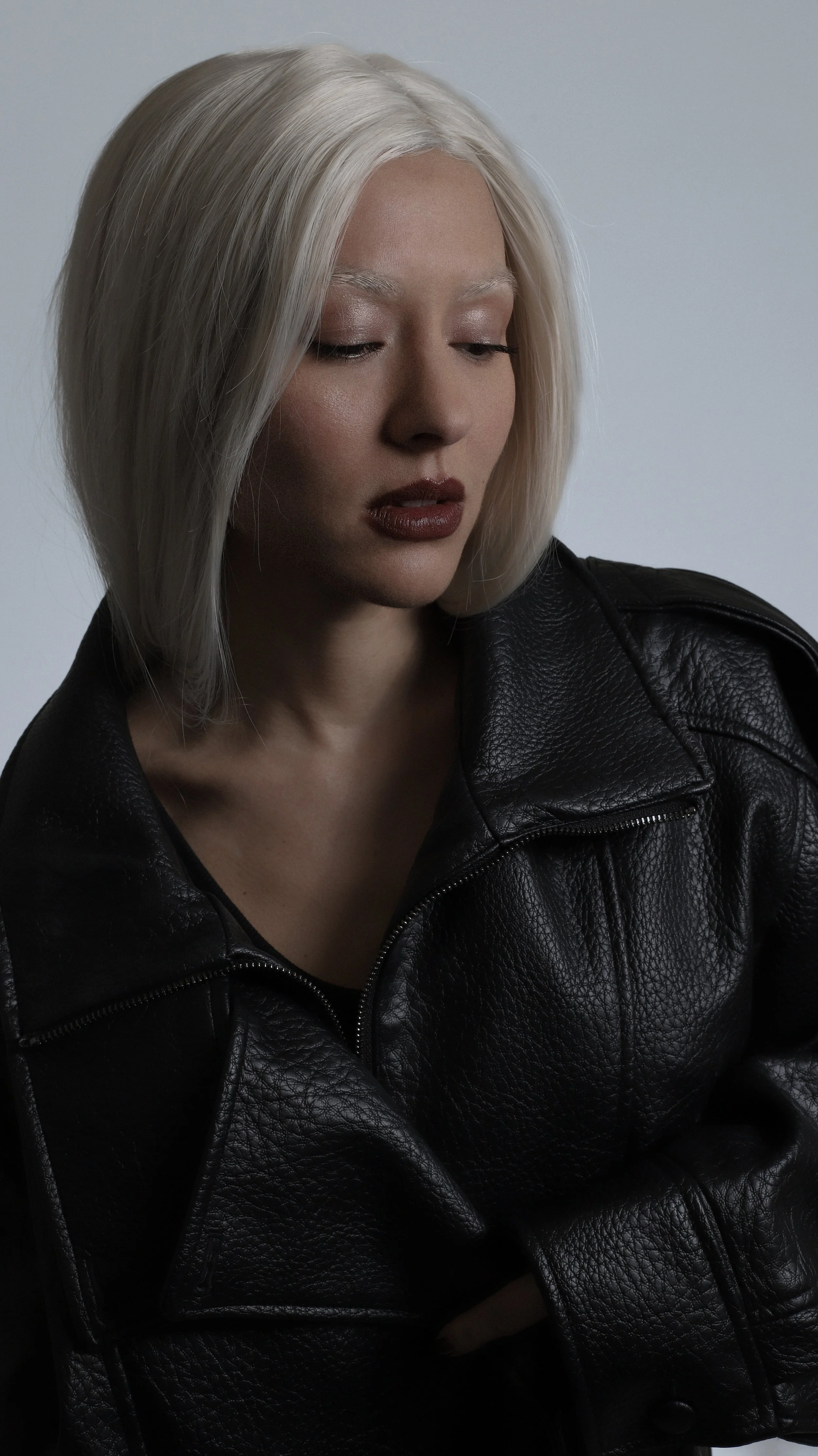 A woman with platinum blonde hair wearing a black leather jacket, looking down with a neutral expression.