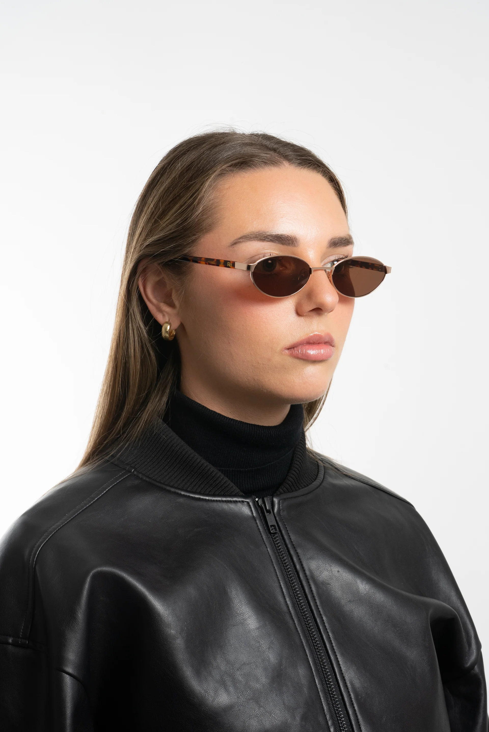 A woman wearing oval sunglasses, black leather jacket, black turtleneck, and gold hoop earrings.