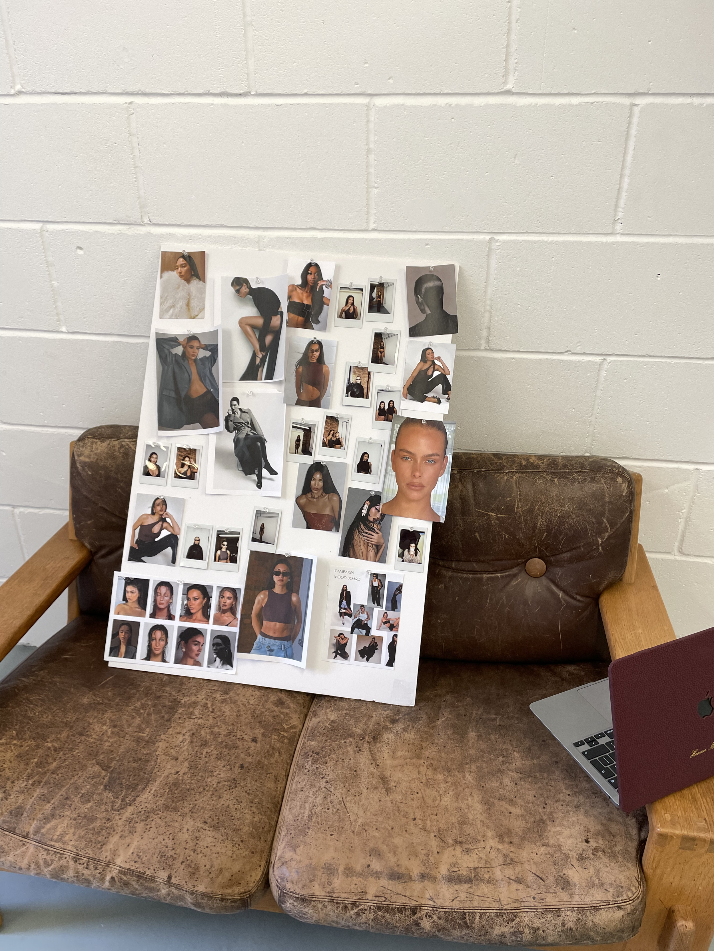 Collage of fashion photos and portraits of models on a white board, placed on a worn brown leather and wood bench, with a closed laptop nearby, against a white brick wall.