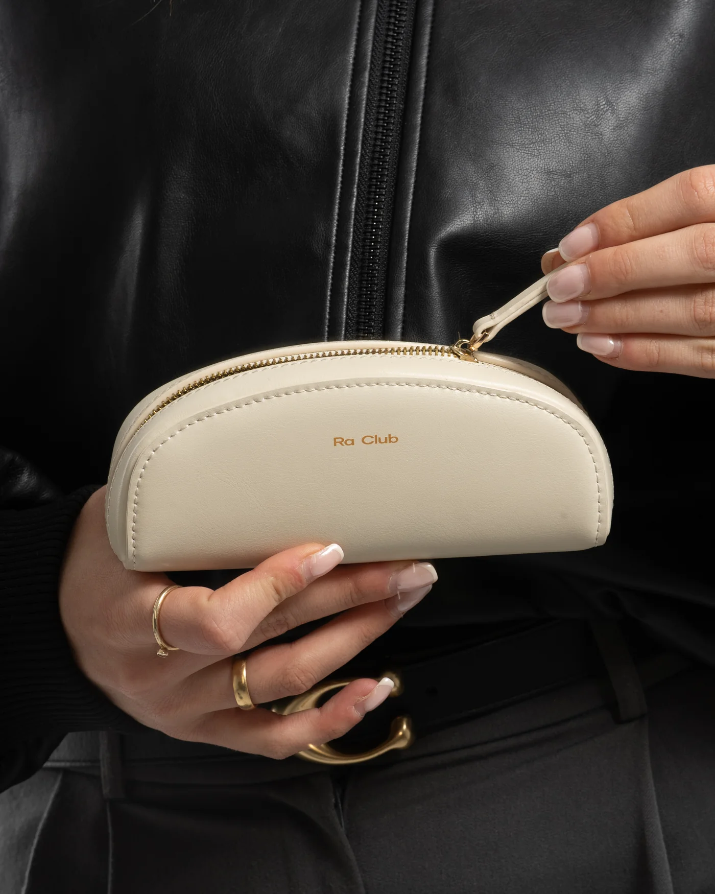 Person holding a cream-colored cosmetic bag with gold zipper, labeled 'Ra Club,' while wearing a black leather jacket and gold rings.