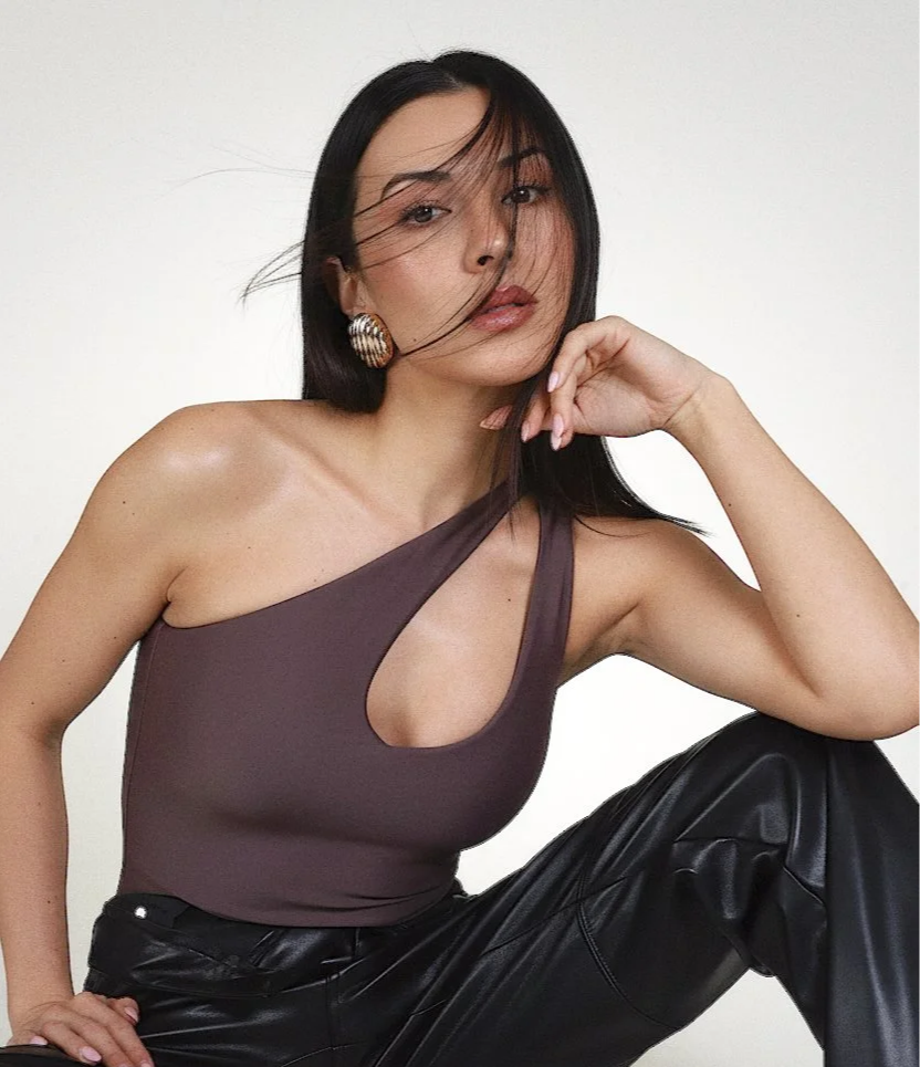 A woman with long dark hair, wearing a one-shoulder brown top with a cutout and black leather pants, posing with one hand near her chin and looking at the camera.