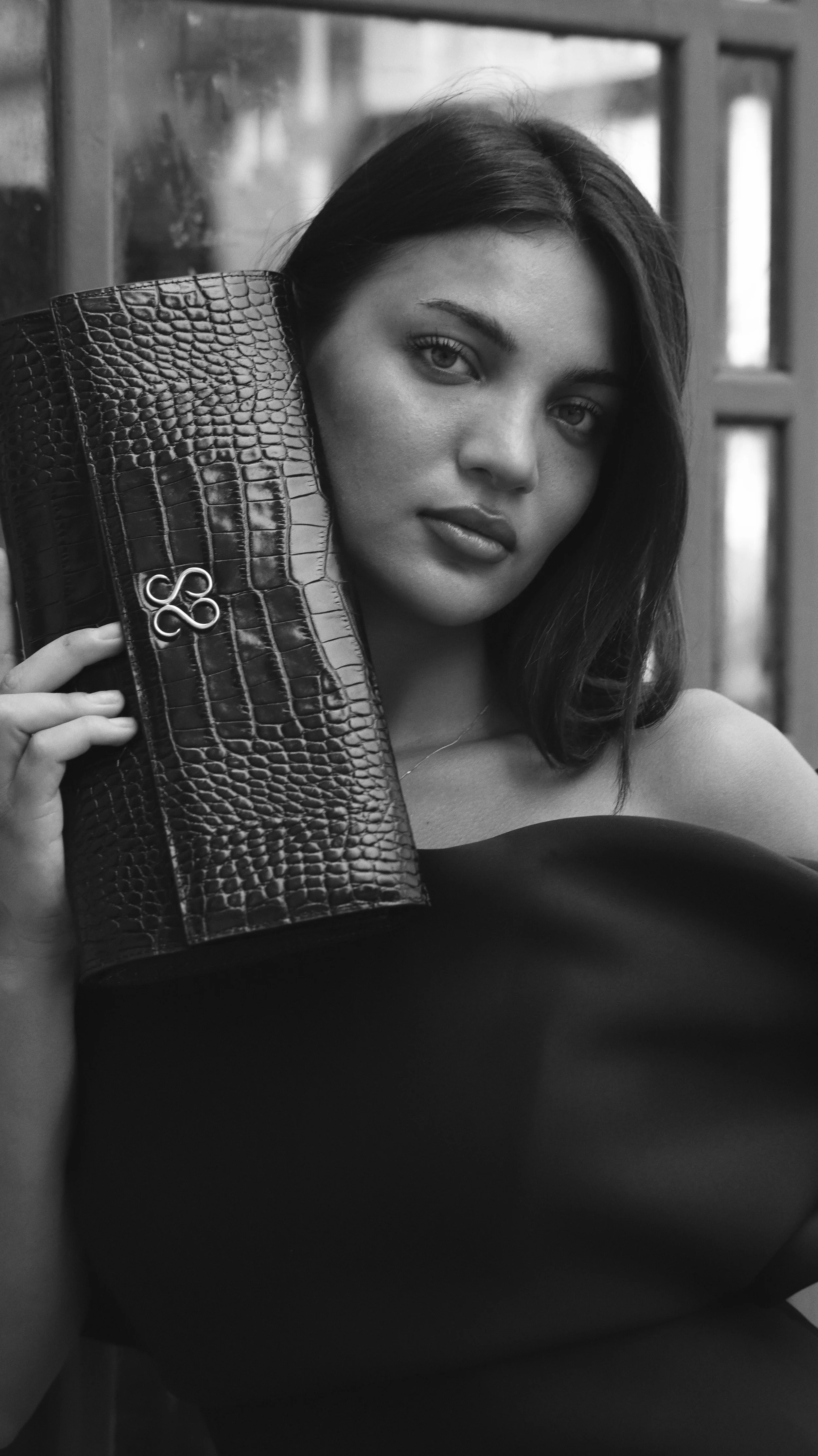 A woman with dark hair and eyes holding a crocodile-skin patterned clutch close to her face, sitting indoors with a window in the background.