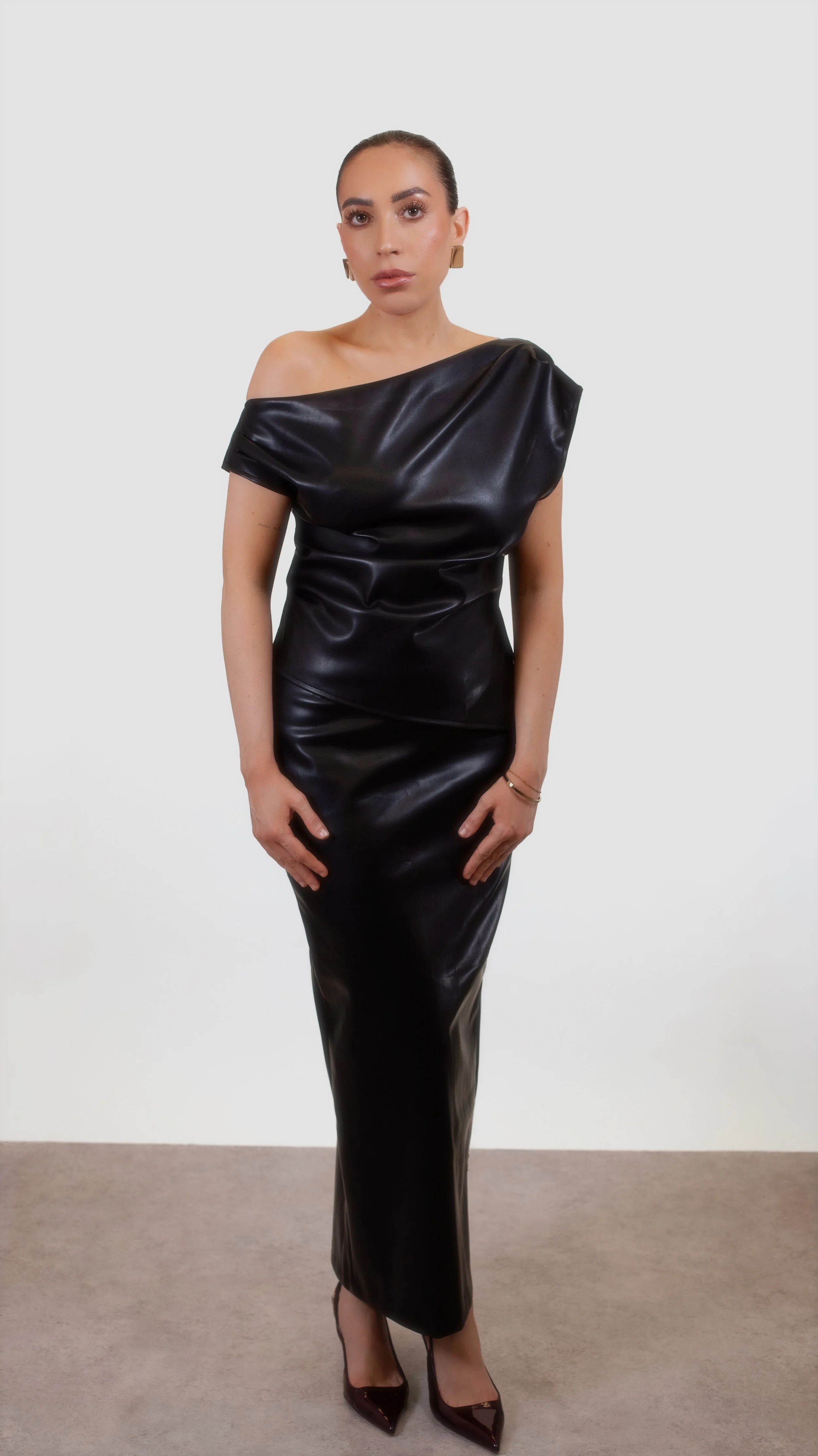 A woman in an off-the-shoulder black leather dress standing against a plain background.