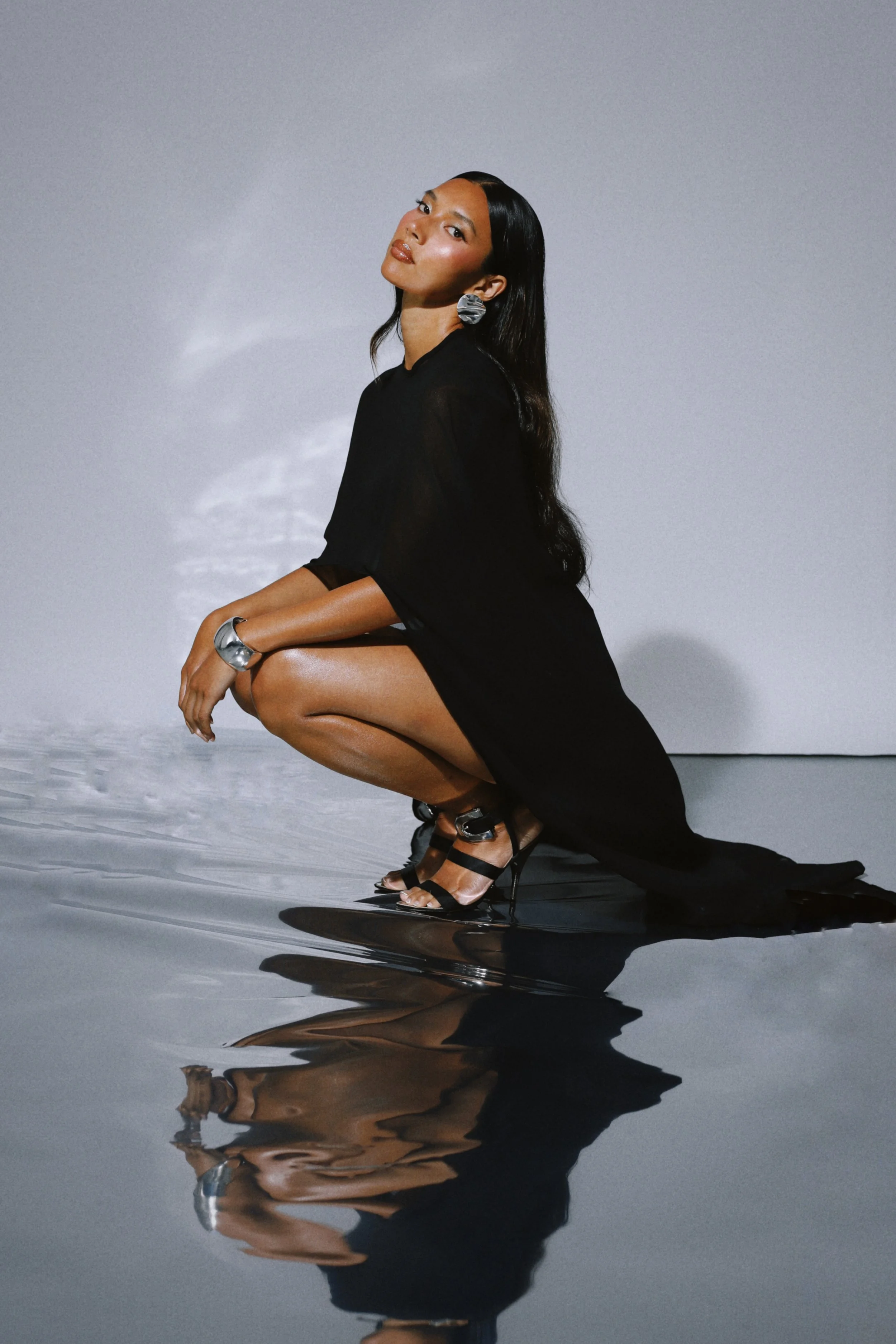 Black sheer kaftan-style dress, test shoot by The Style Strategist, model crouched on silver reflective floor.