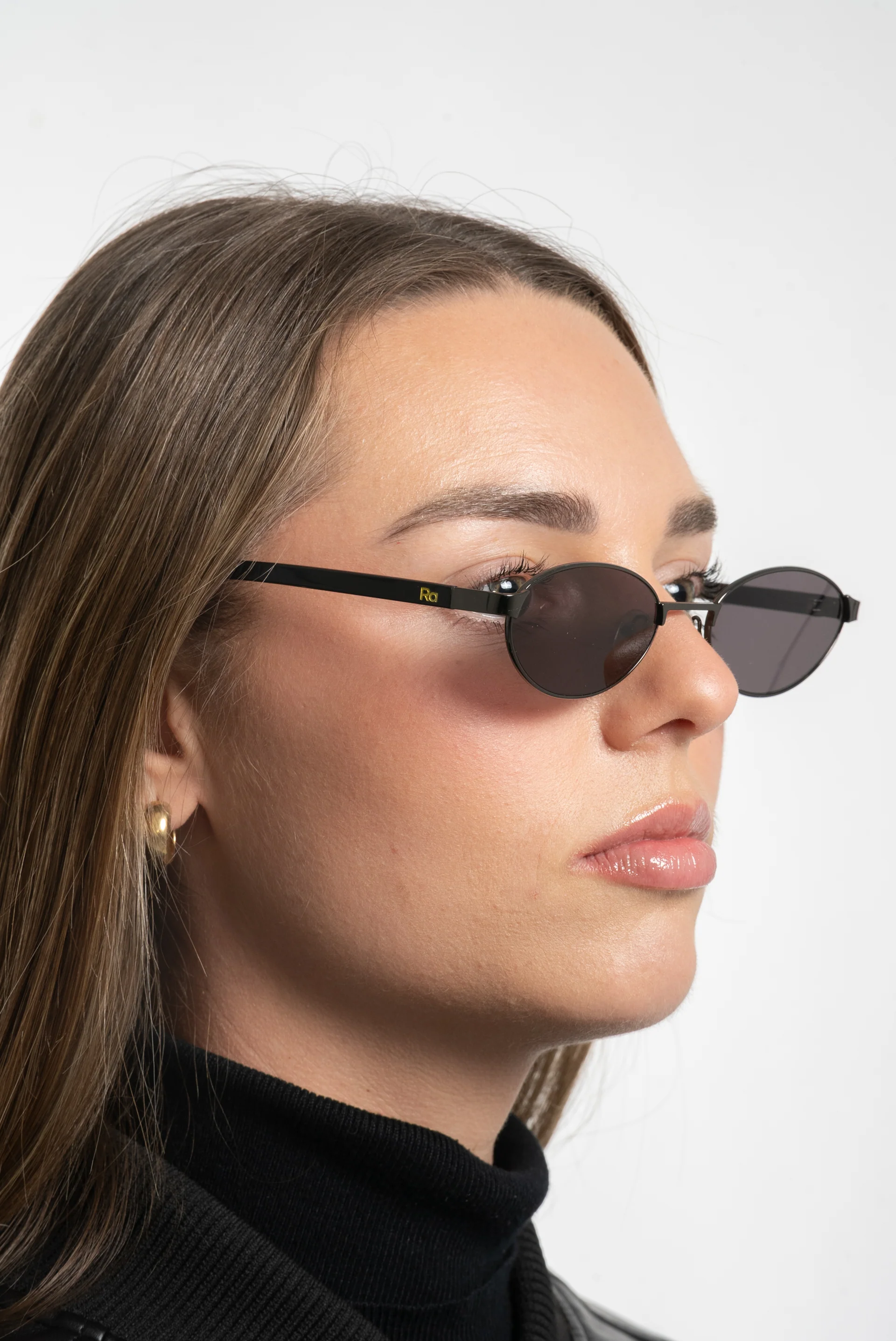 Close-up of a woman with long brown hair wearing black oval sunglasses, gold earrings, and a black turtleneck against a white background.
