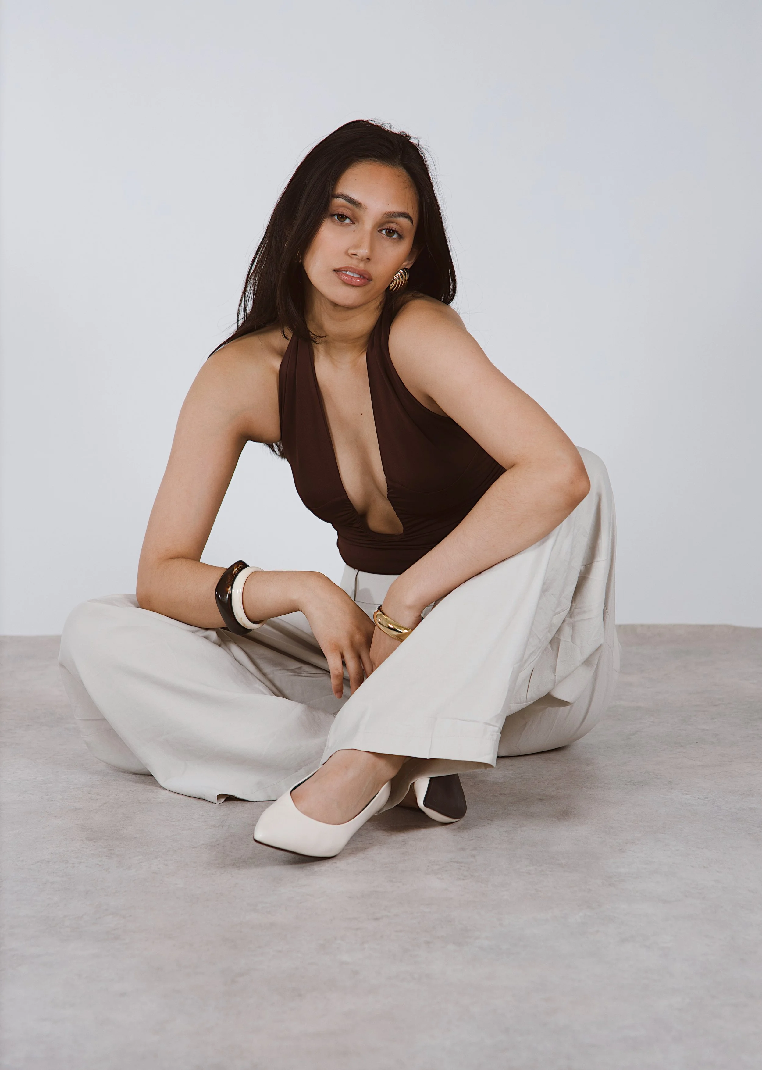 Young woman with long dark hair, wearing a sleeveless brown top and beige wide-leg pants, sitting on the floor with one leg crossed and the other bent, posing against a plain light background.