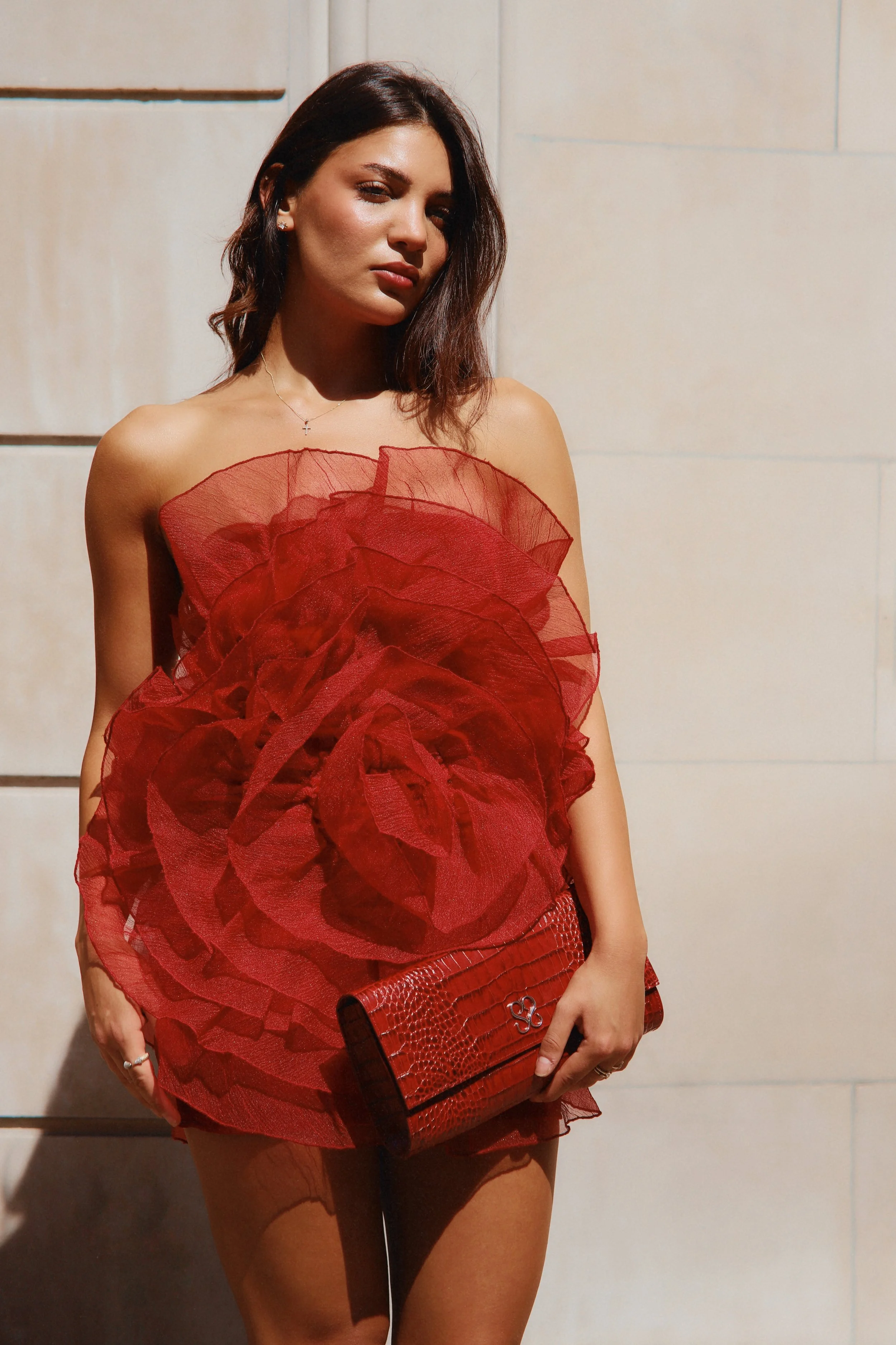 Woman with dark brown hair wearing a red ruffled dress, holding a red clutch, standing against a light-colored wall.