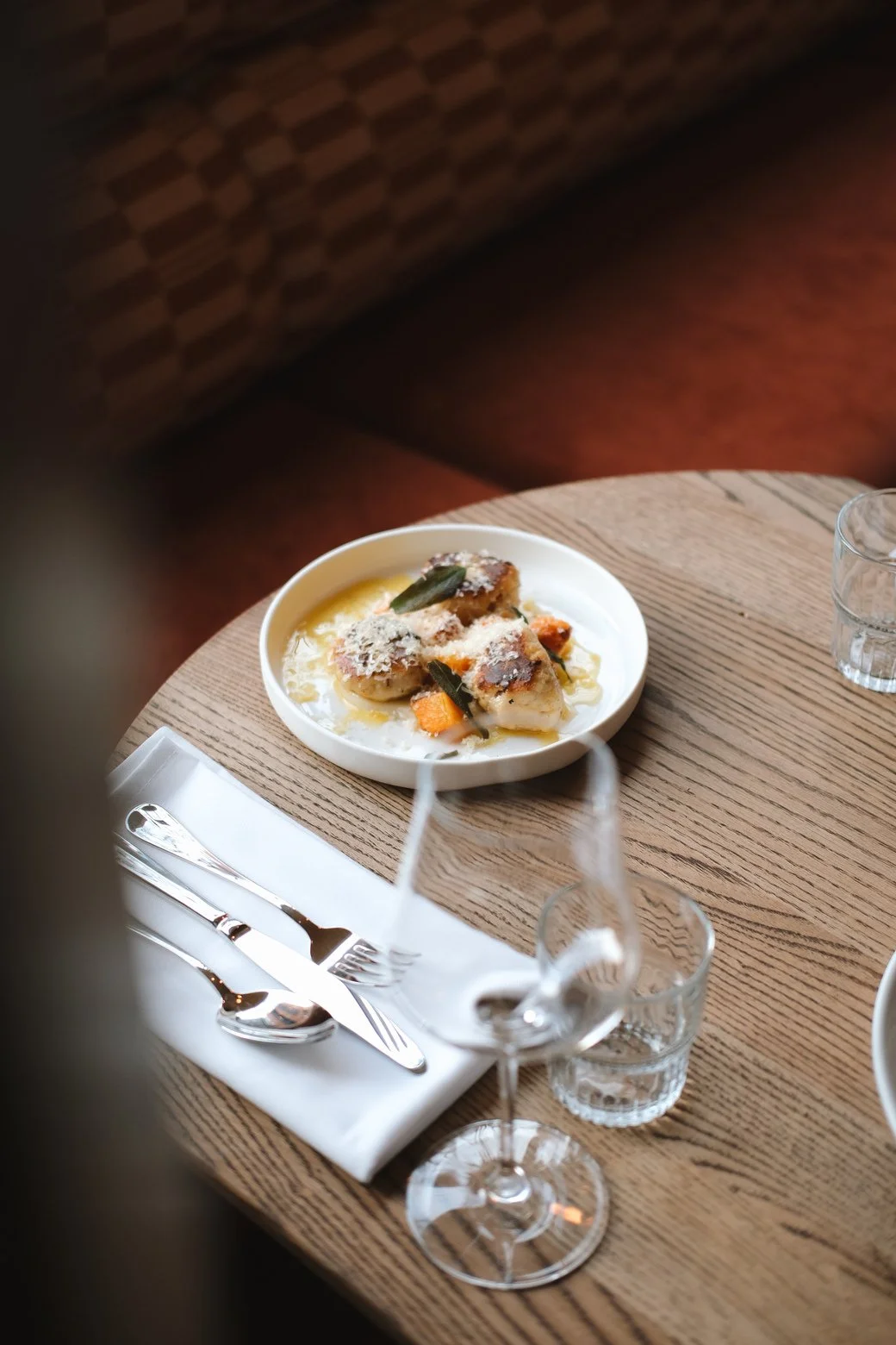 A plate of food with fish, vegetables, and sauce on a wooden table, accompanied by utensils, wine glass, and water glasses.