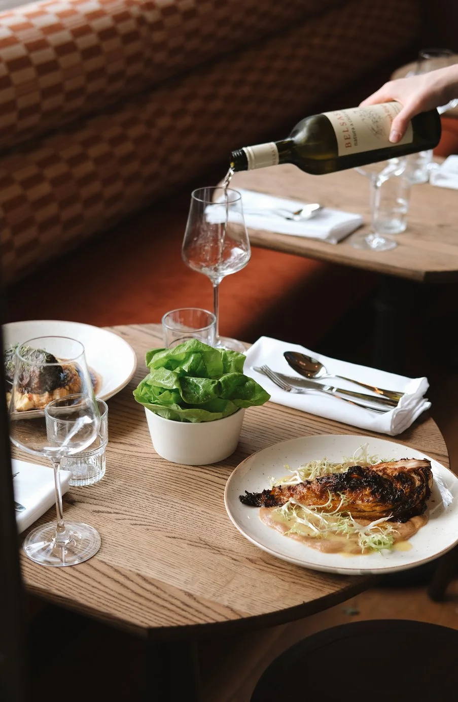 A restaurant scene with a wooden table, a white plate with a roasted chicken drumstick, a small bowl of green salad, a wine glass being filled with white wine, and additional glasses and plates set for dining.