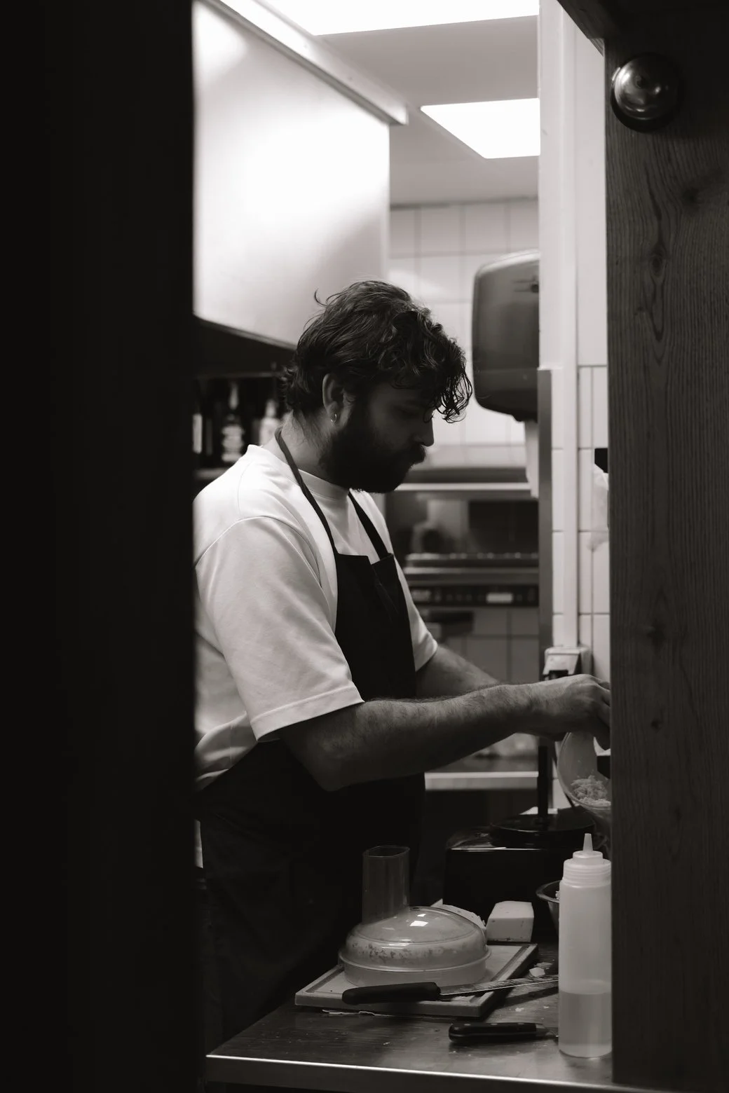 Black and white photo of a man in a kitchen, preparing food, with a beard and curly hair, wearing an apron and a white t-shirt.