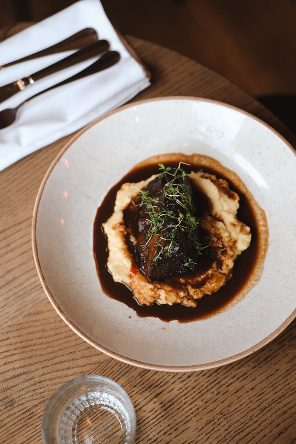 A plate of mashed potatoes topped with a braised beef short rib, garnished with microgreens, served with dark gravy on a white ceramic dish.