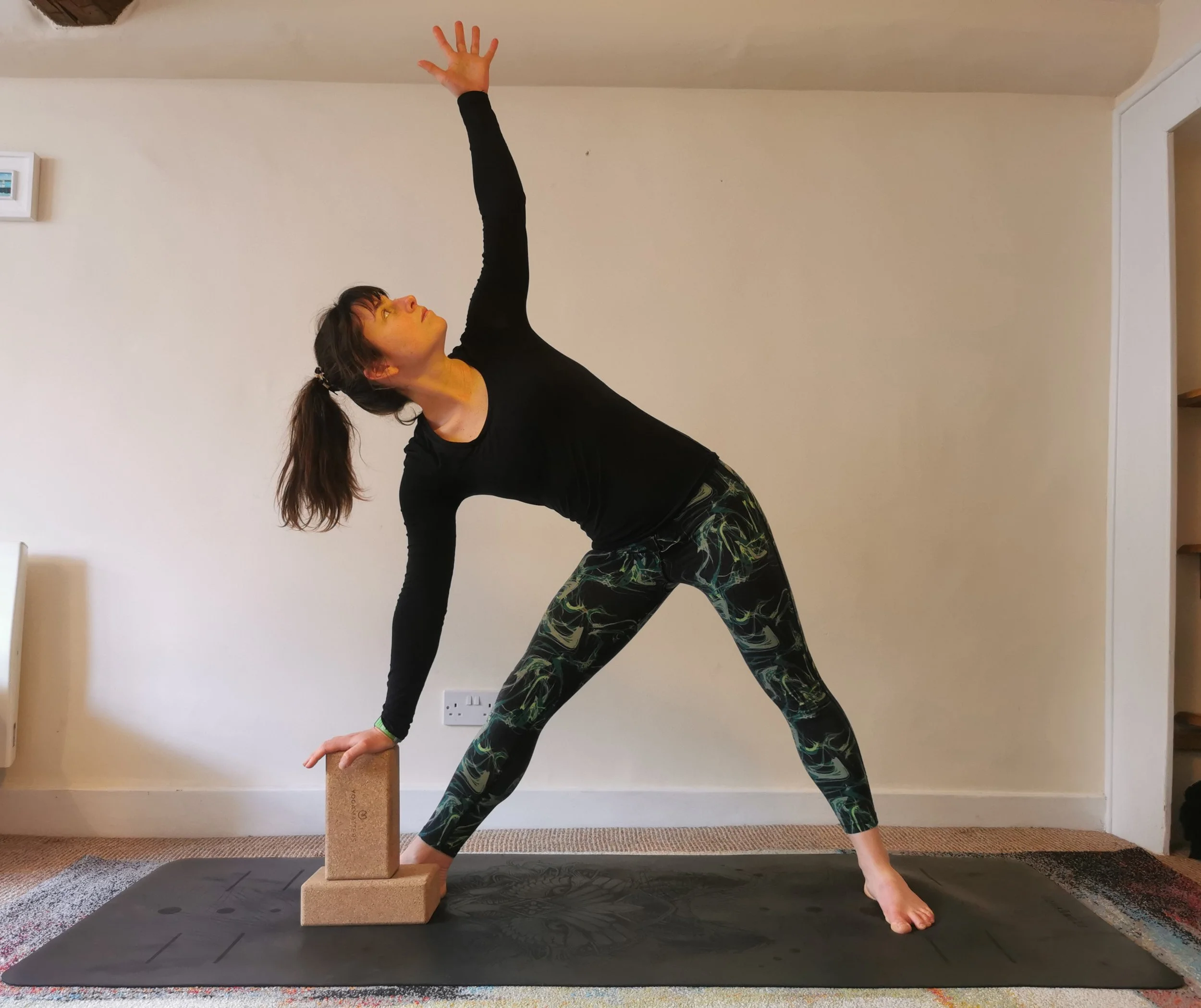 A woman practicing yoga indoors, in warrior pose with one arm reaching up and the other hand on a yoga block, on a black yoga mat.