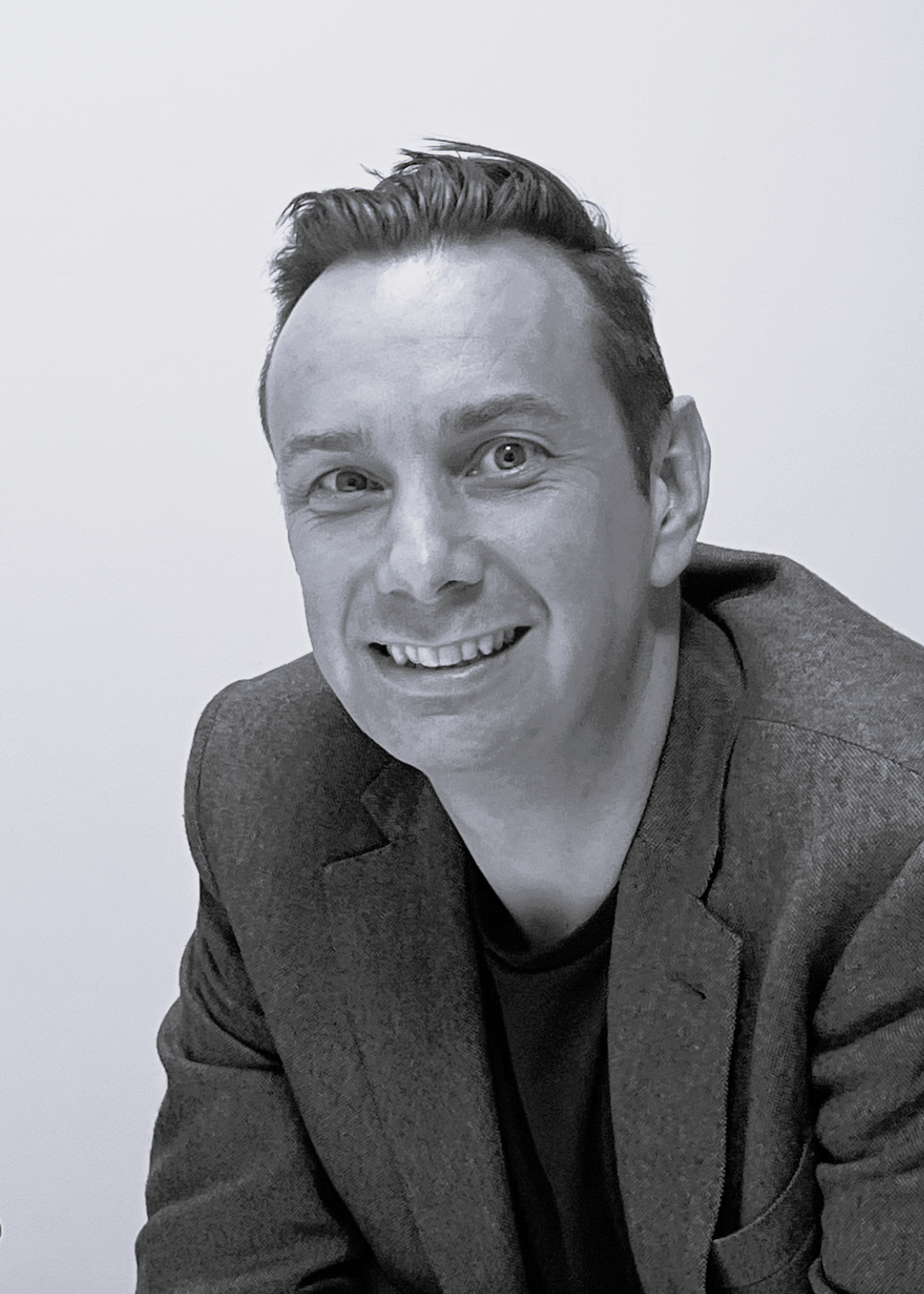 A black and white photo of a man with short, dark hair wearing a blazer, smiling and leaning slightly forward.