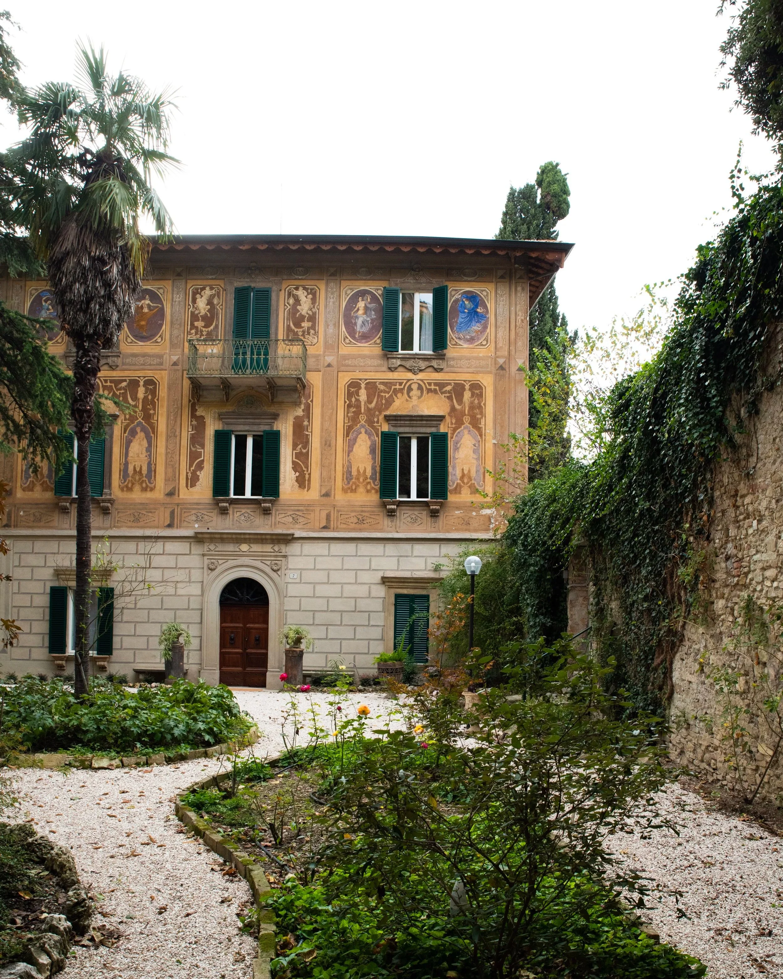 Edificio storico con affreschi e finestre con tapparelle verdi, giardino con piante e sentieri di ghiaia