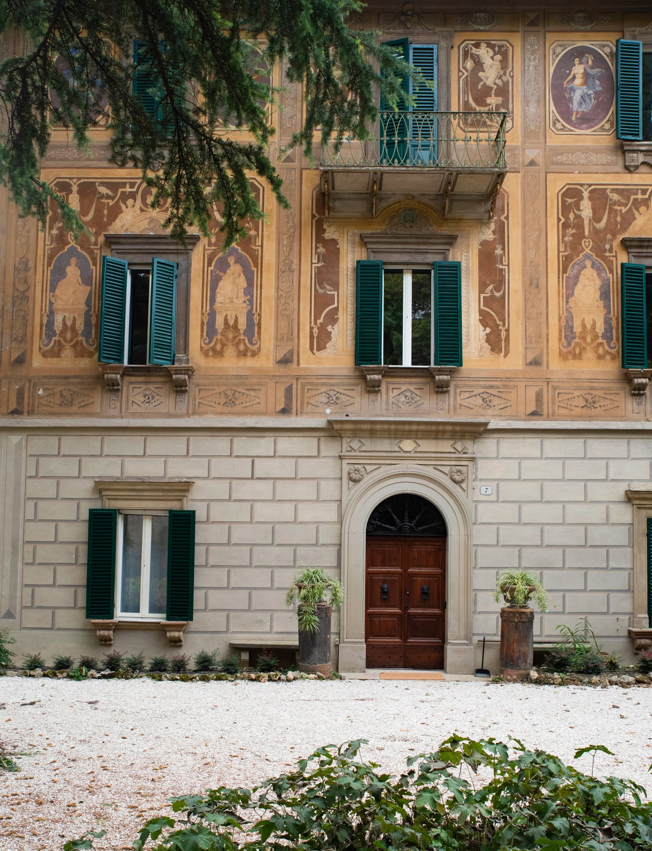 Facciata di un palazzo storico con finestre con persiane verdi, un portone in legno, decori sulle pareti e un balcone con ringhiera verde. Ci sono piante ai lati dell'ingresso e un albero che copre parzialmente la parte superiore dell'immagine.