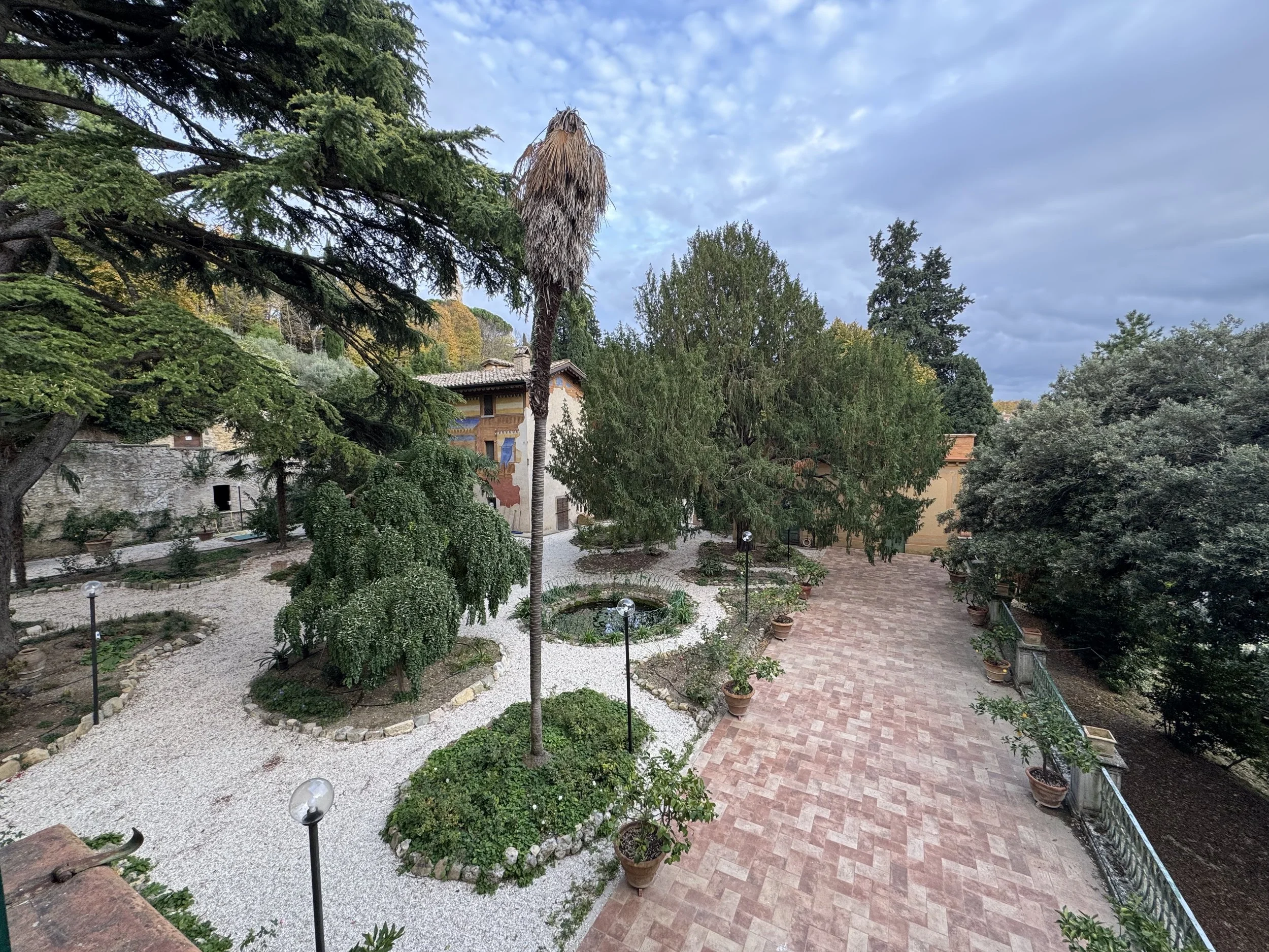 Giardino con sentieri di ghiaia e piante in vaso, grandi alberi e un edificio in lontananza, sotto un cielo nuvoloso.