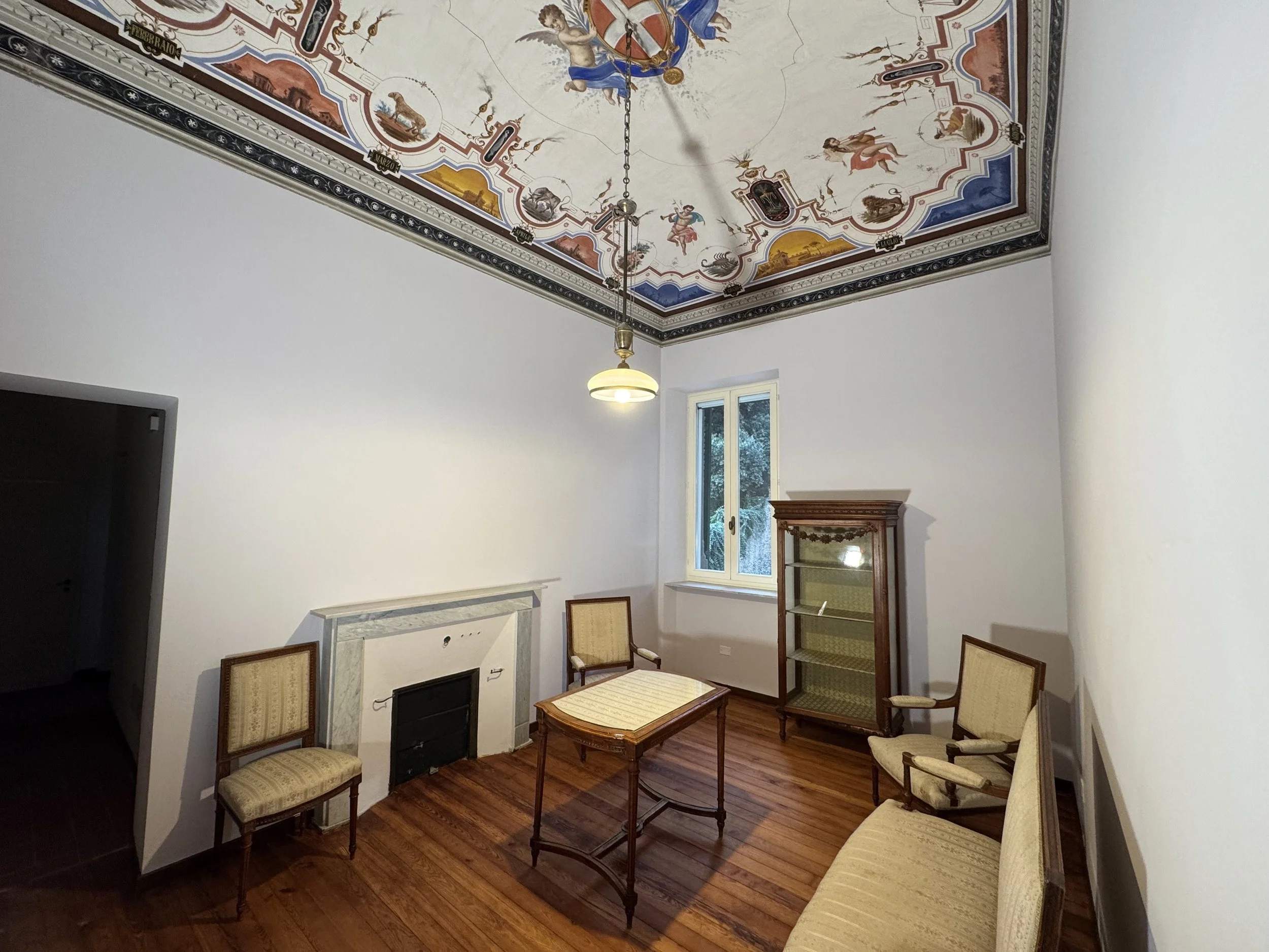 Stanza con soffitto affrescato con figure zodiacali, pareti bianche, camino bianco, finestrella e mobili d'antiquariato, tra cui sedie, tavolo e vetrina in legno.