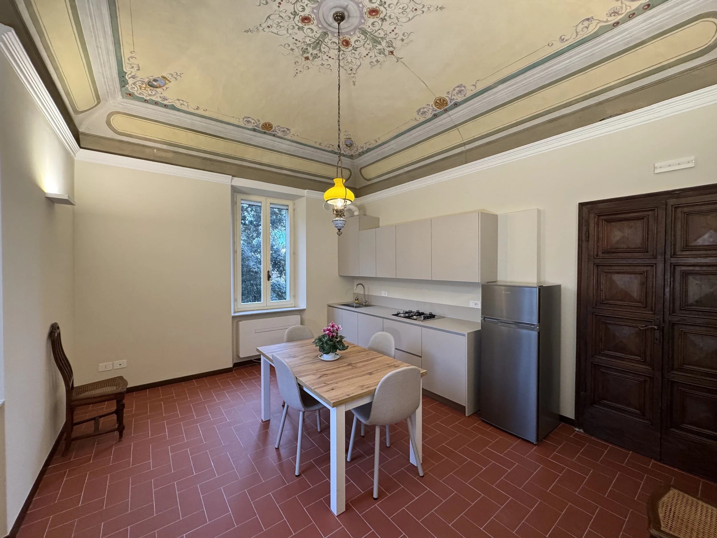 Cucina moderna con soffitto decorato, tavolo in legno con quattro sedie, frigorifero in acciaio, armadio in legno scuro, finestra con vista sugli alberi, lavabo, coclee e armadietti bianchi.