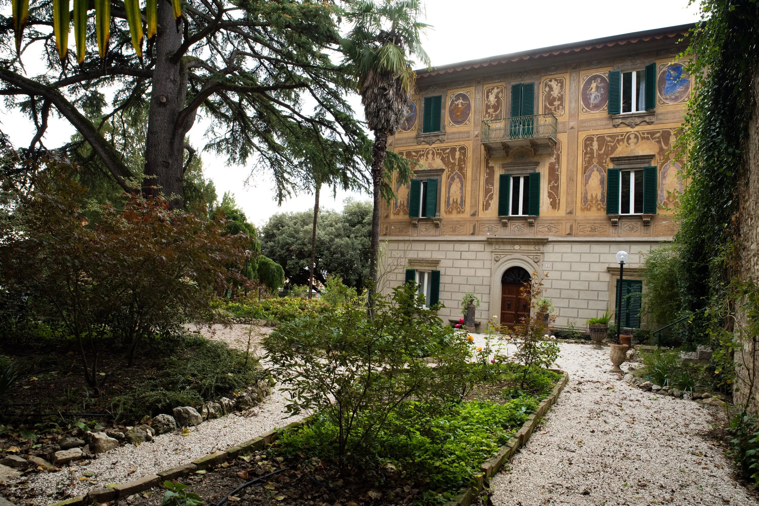 Giardino con piante e fiori di fronte a un palazzo storico con pareti decorate e finestre con persiane verdi.