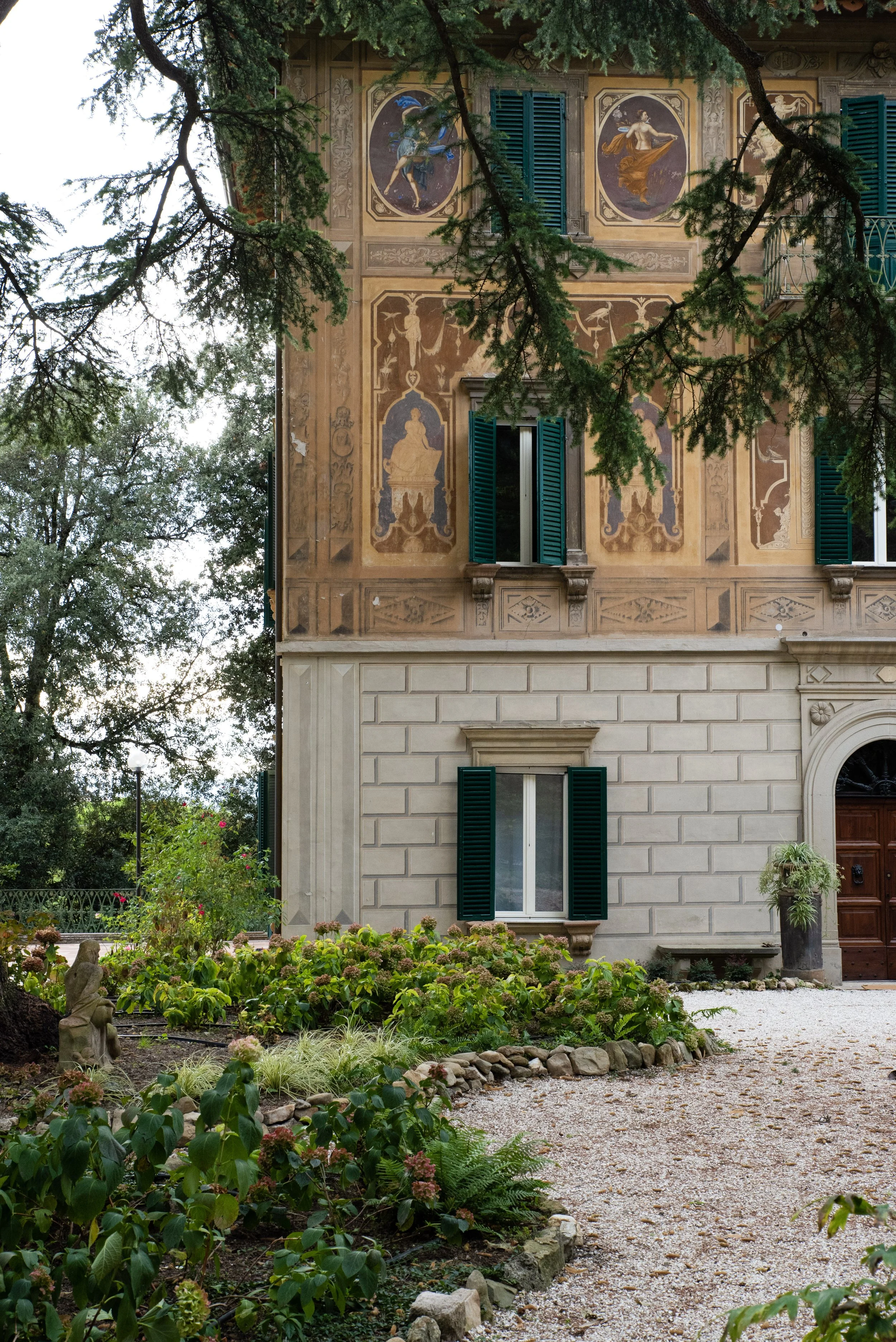 Edificio storico con affreschi sulla facciata, finestra con imposte verdi, giardino con piante e statue, strada sterrata.