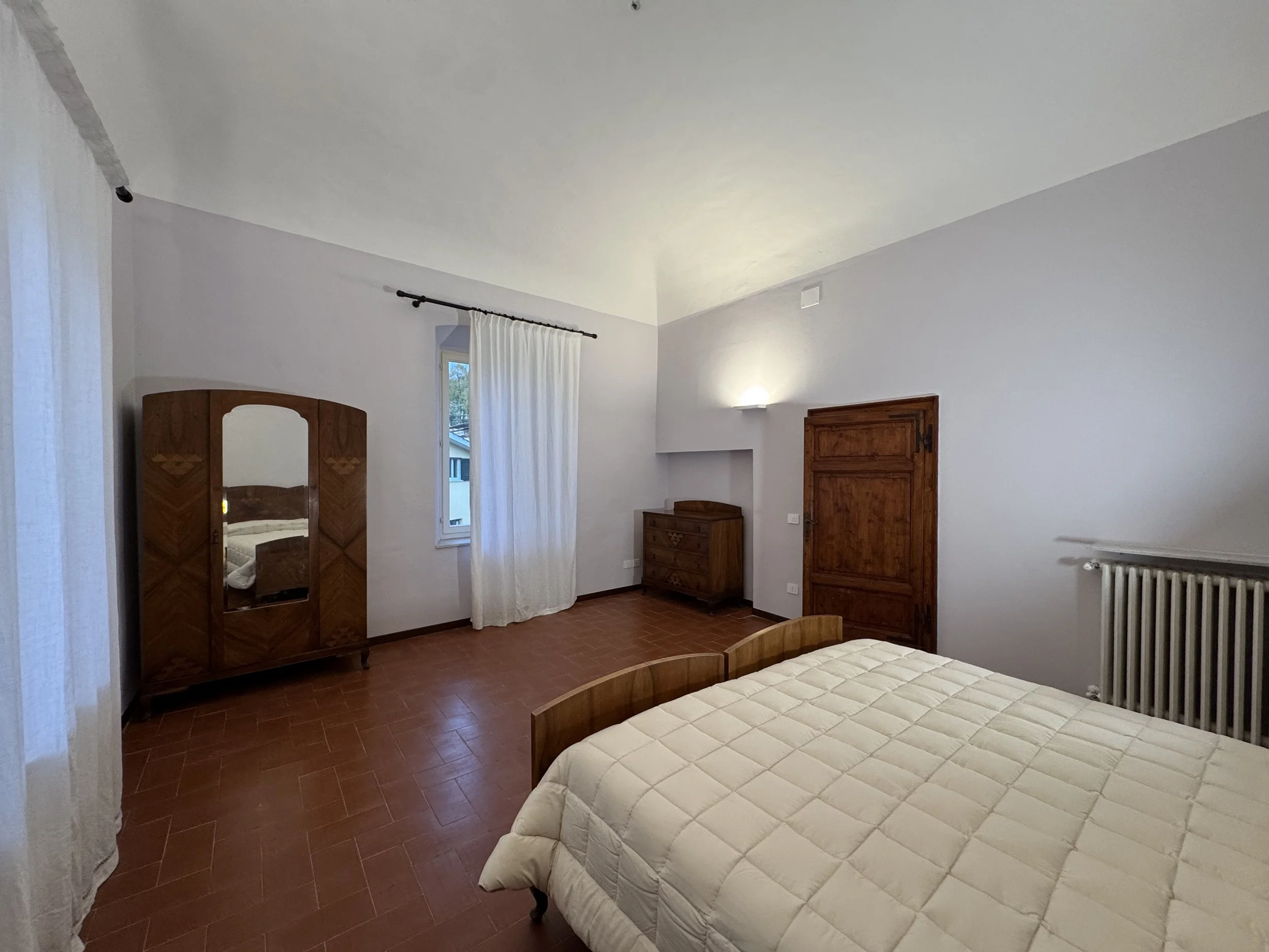 Camera da letto con un letto matrimoniale, armadio con specchio, davanzale con finestra, cassettiera in legno, portasfortuna, termosifone e pareti bianche.