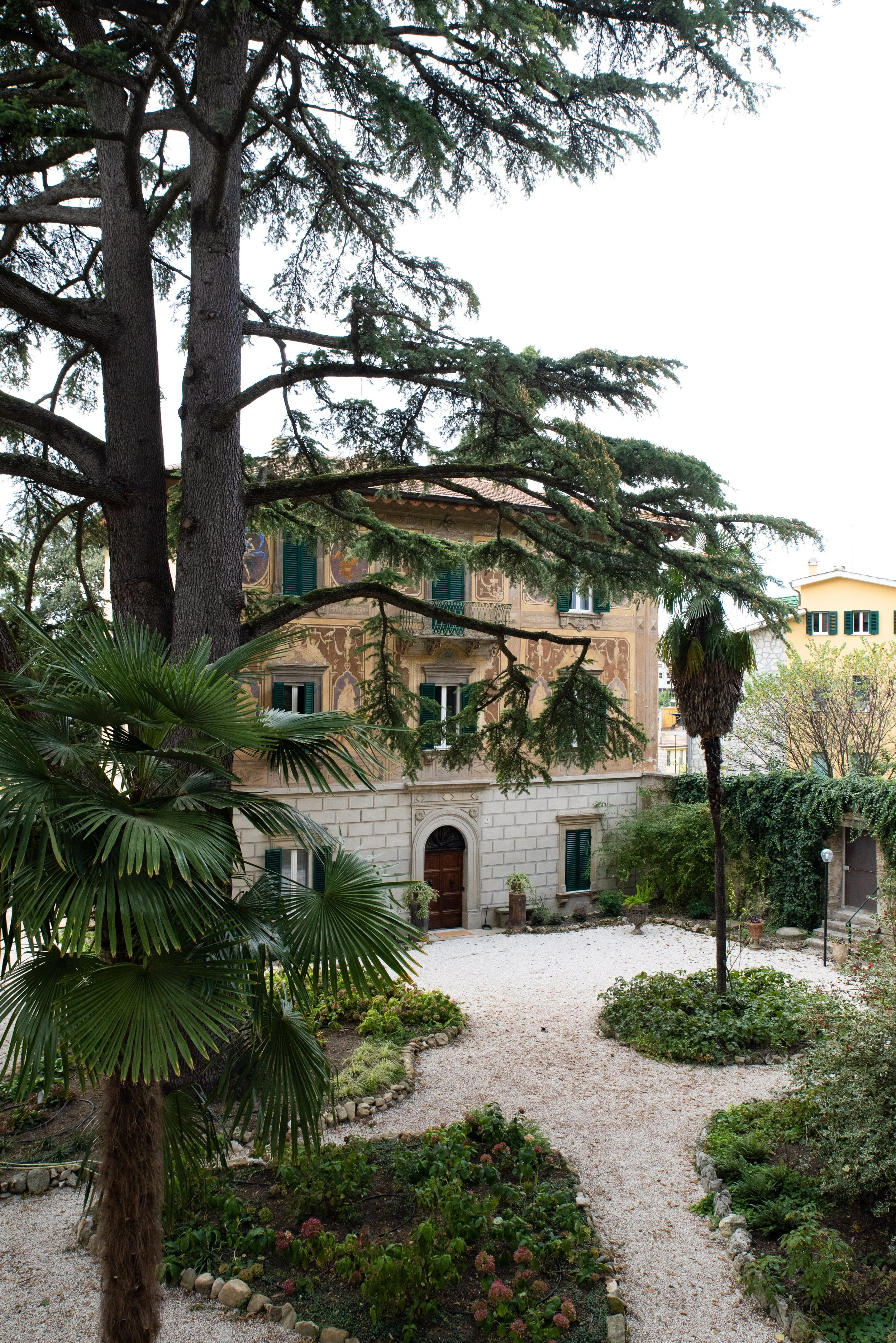 Giardino con piante verdi, un grande albero e un edificio antico sullo sfondo.