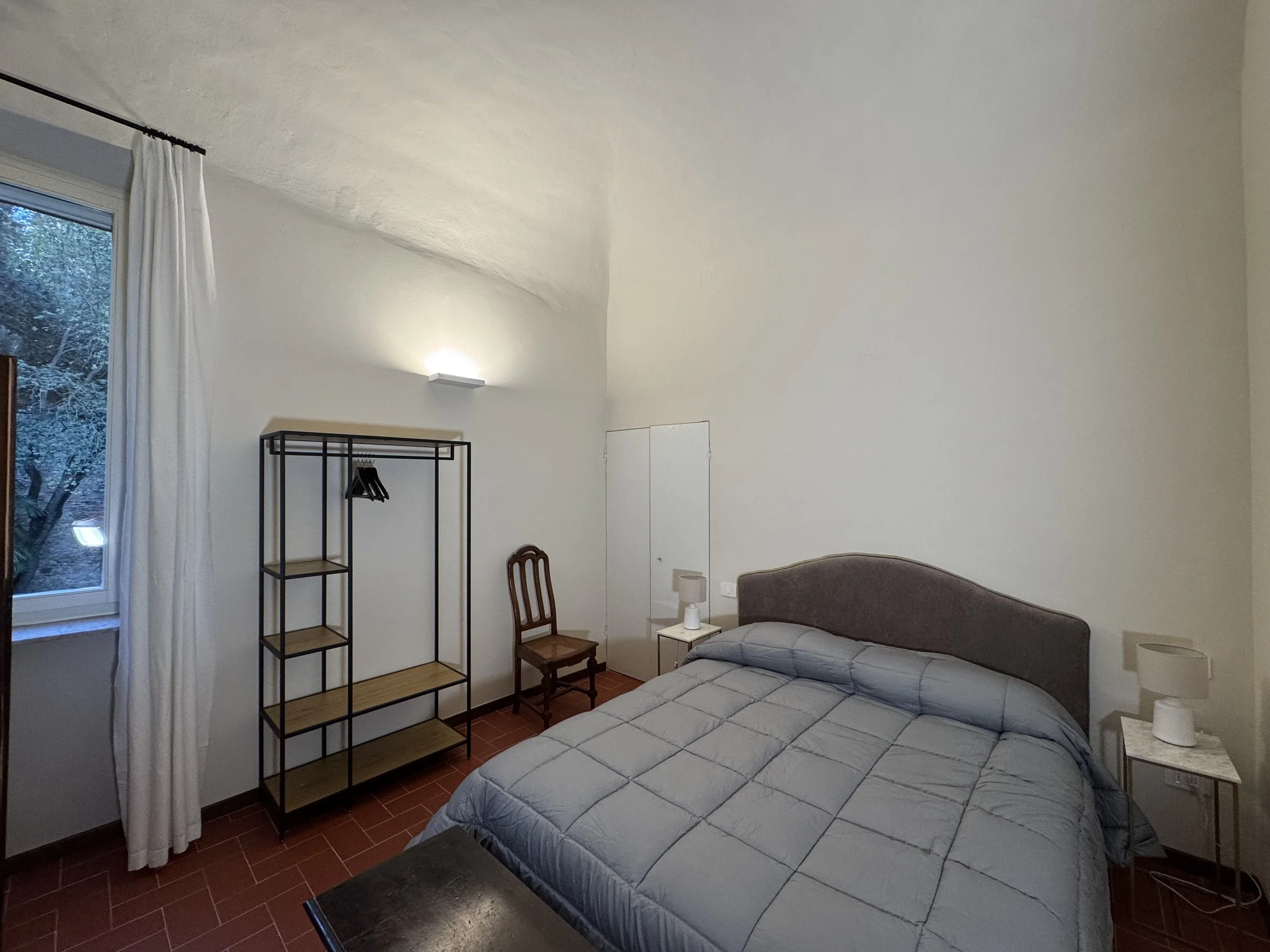 Camera matrimoniale con letto, due lampade da tavolo, sedia in legno, scaffale con guardaroba e finestra con tende bianche.