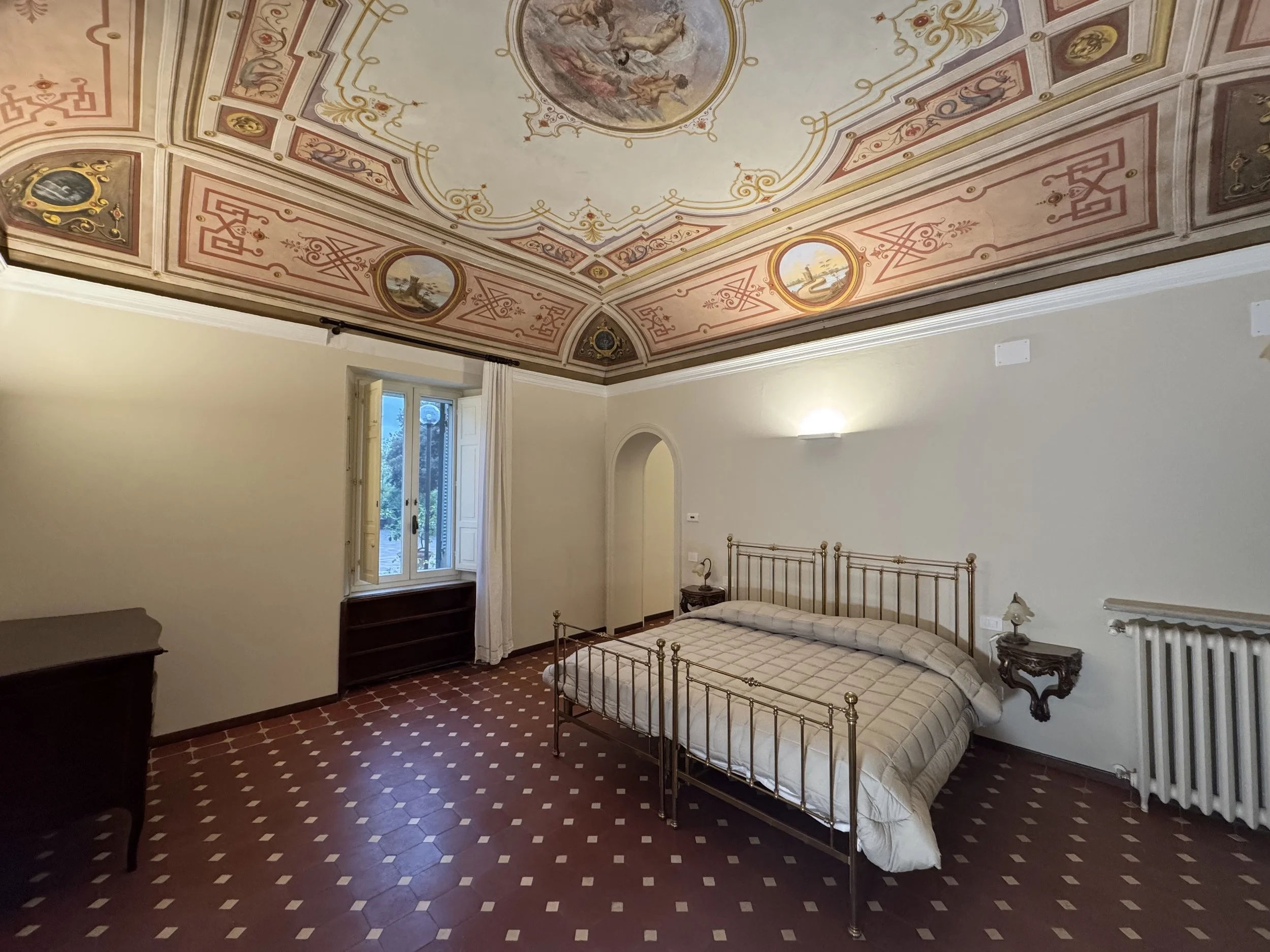 Camera da letto con soffitto affrescato, letto in metallo battuto, due comodini con lampade, finestra con tende bianche, termosifone, pavimento di piastrelle rosse e bianche.