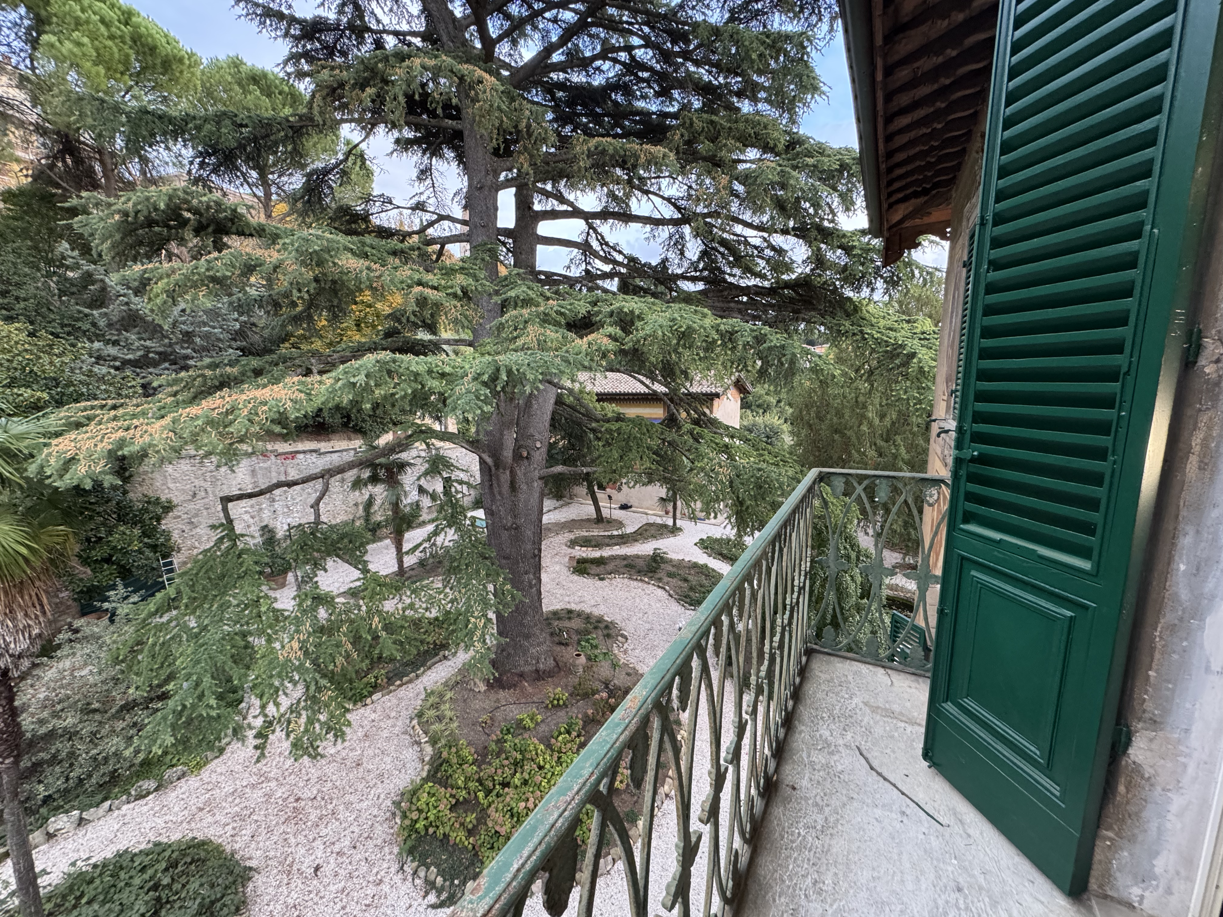 Vista dal balcone con grate in ferro battuto e porta con persiana verde, che si affaccia su un giardino con alberi e cortile di pietra.