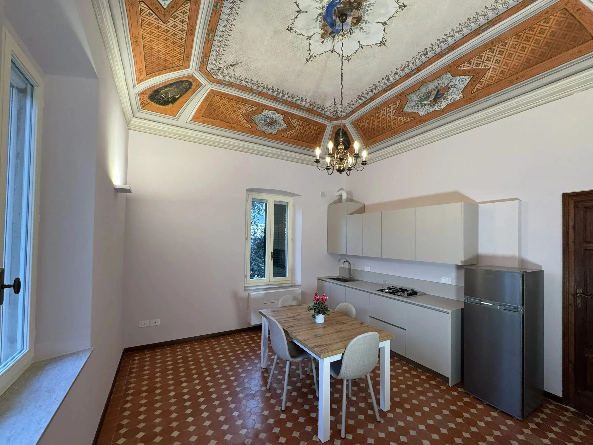 Interno di una cucina con soffitto decorato, pareti bianche, mobili chiari, tavolo in legno con quattro sedie, frigorifero in acciaio, lavabo, finestra e porta in legno.