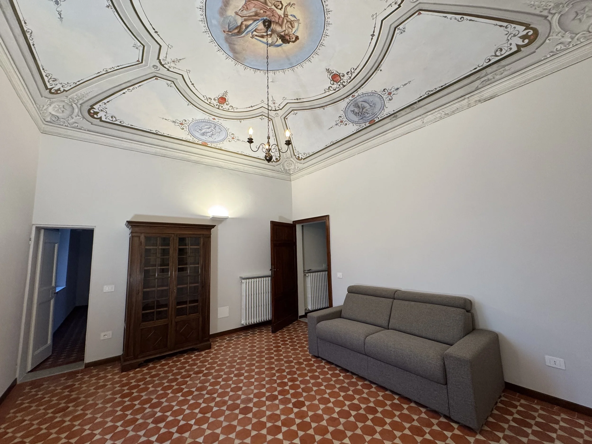 Stanza con soffitto decorato con dipinto e decorazioni, poltrona grigia, mobile in legno, porte in legno e pavimento con piastrelle a mosaico.