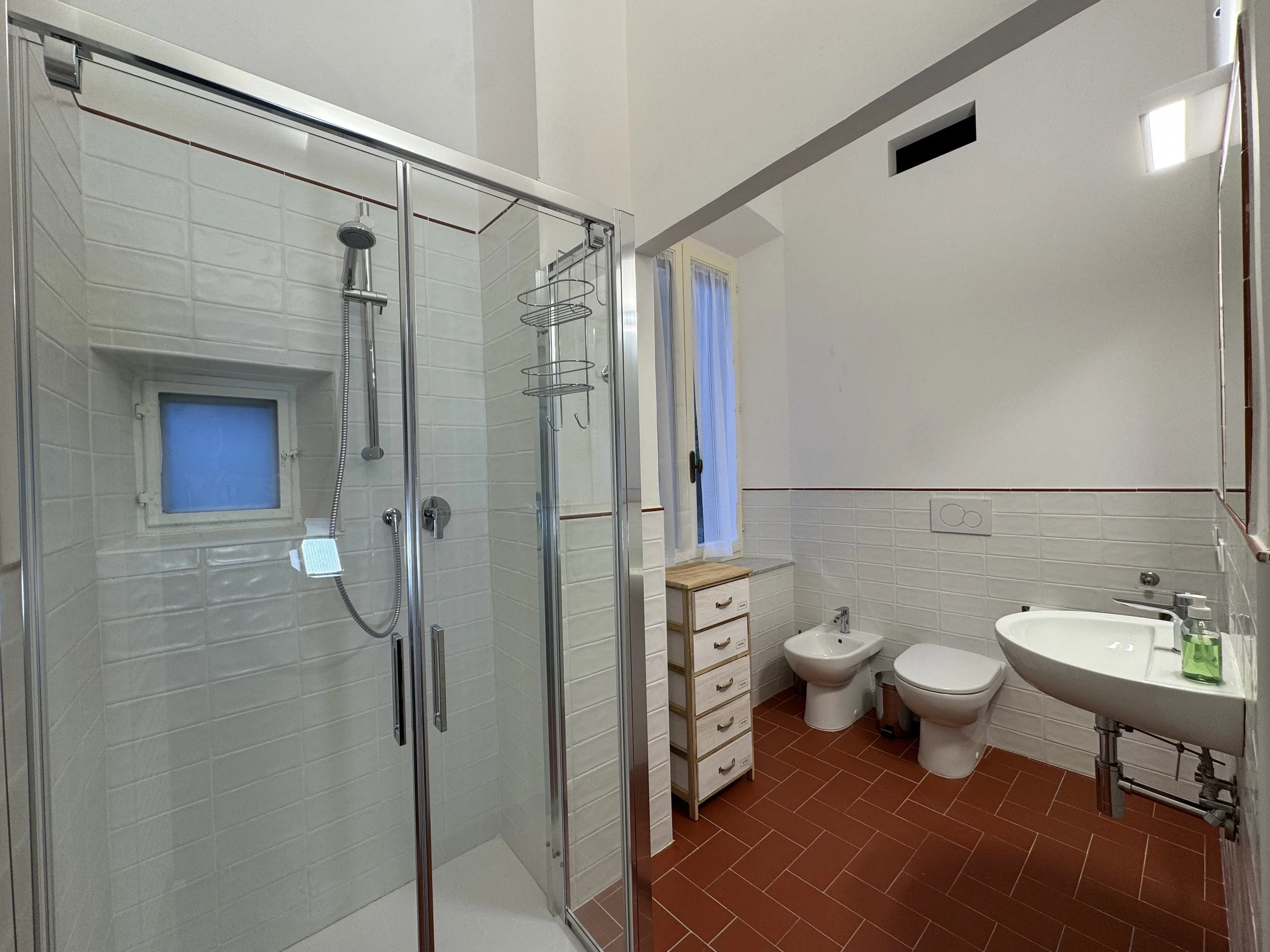Bagno con cabina doccia, bidet, toilet, lavabo, finestra, pareti rivestite di mattoni bianchi, pavimento in piastrelle rosse