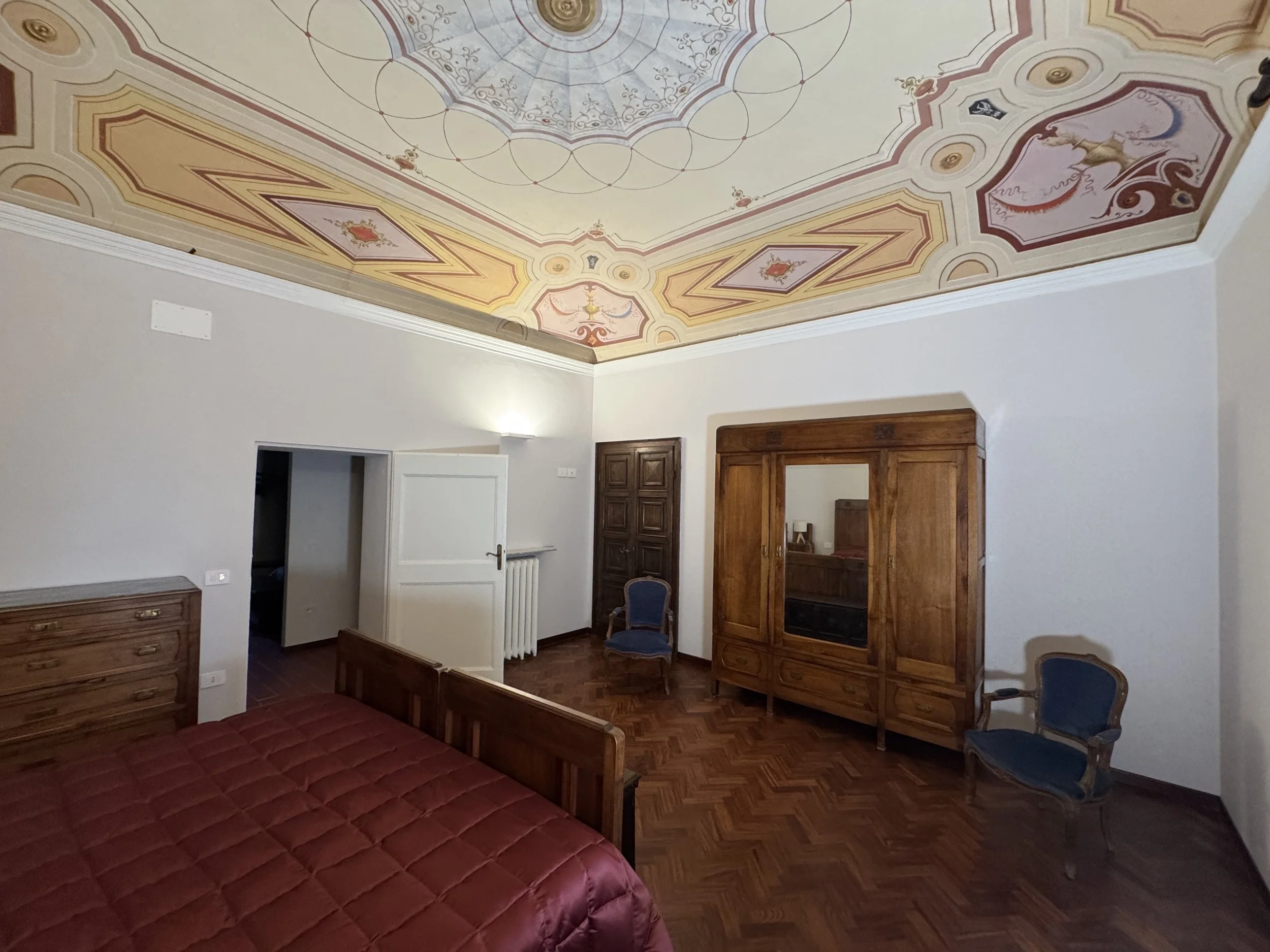 Camera da letto con soffitto affrescato, letto con copriletto cremisi, armadio in legno, due sedie blu in stile antico, pareti bianche, pavimento in parquet.