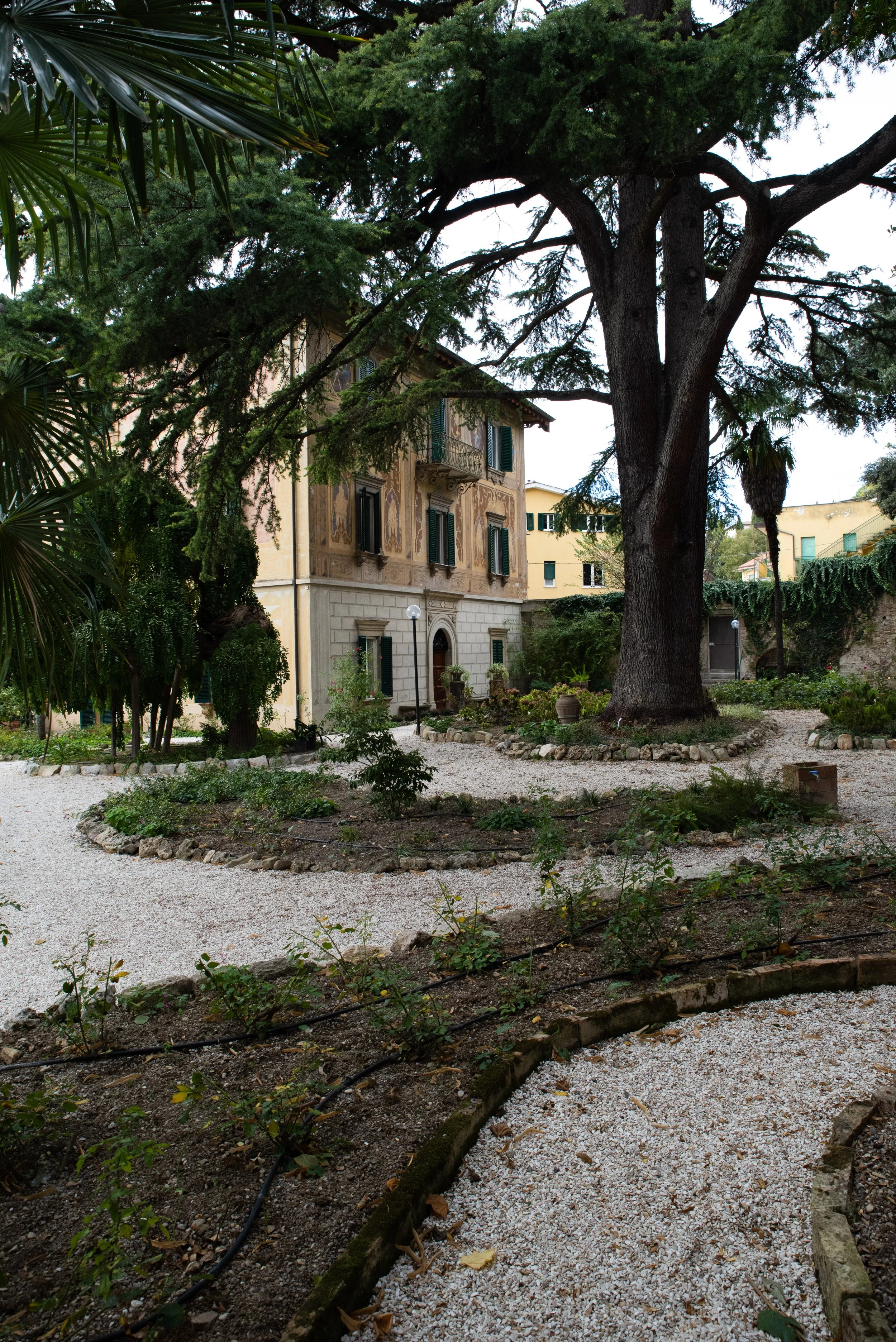 Giardino con grandi alberi e sentieri di ghiaia davanti a una casa storica con finestre con persiane verdi.