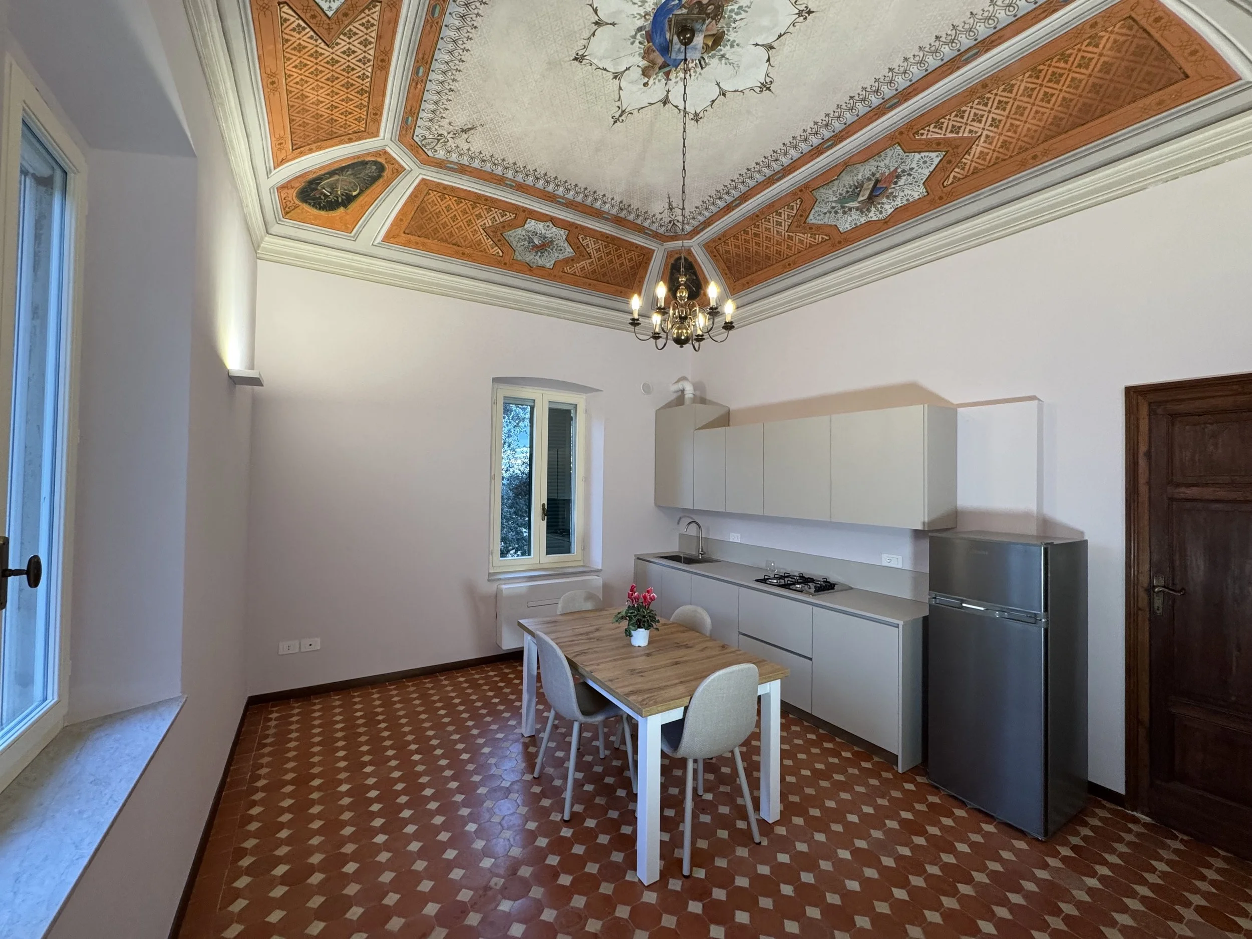 Una cucina moderna in una stanza con pareti bianche, pavimento damier di terracotta, e un soffitto decorato con affreschi e un lampadario color oro. Include una tavola in legno con quattro sedie, un frigorifero in acciaio e armadietti beige sopra il 