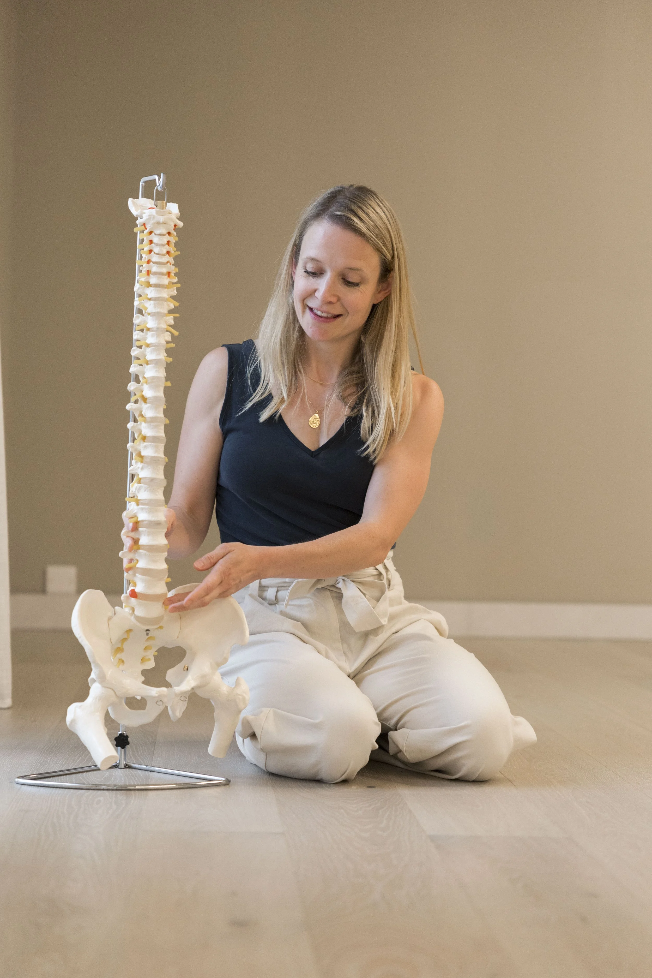 A woman sitting on the floor next to a life-sized anatomical model of a human spine and pelvis, pointing at the pelvis, in a neutral-colored room.