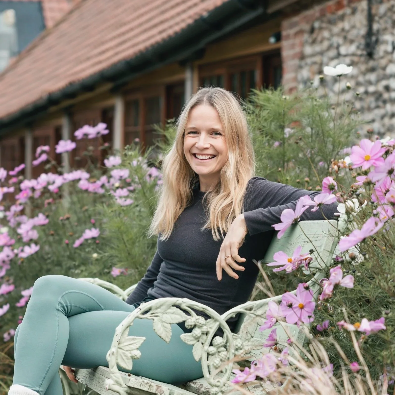 A young woman with blonde hair and light skin sitting on a decorative white metal garden bench surrounded by pink flowers, smiling at the camera.