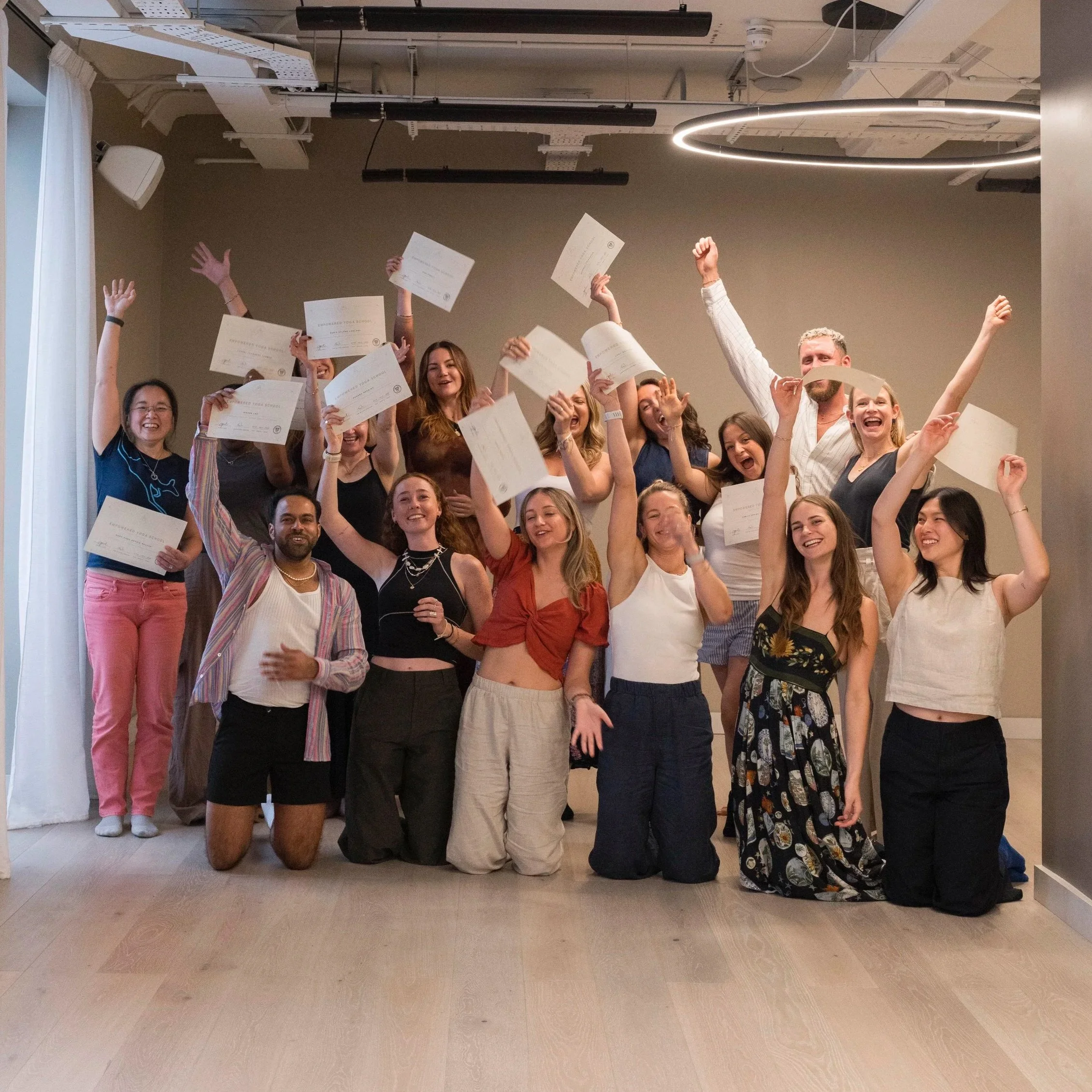 Group of young adults celebrating with certificates, some are kneeling, some are standing, with arms raised and smiling.